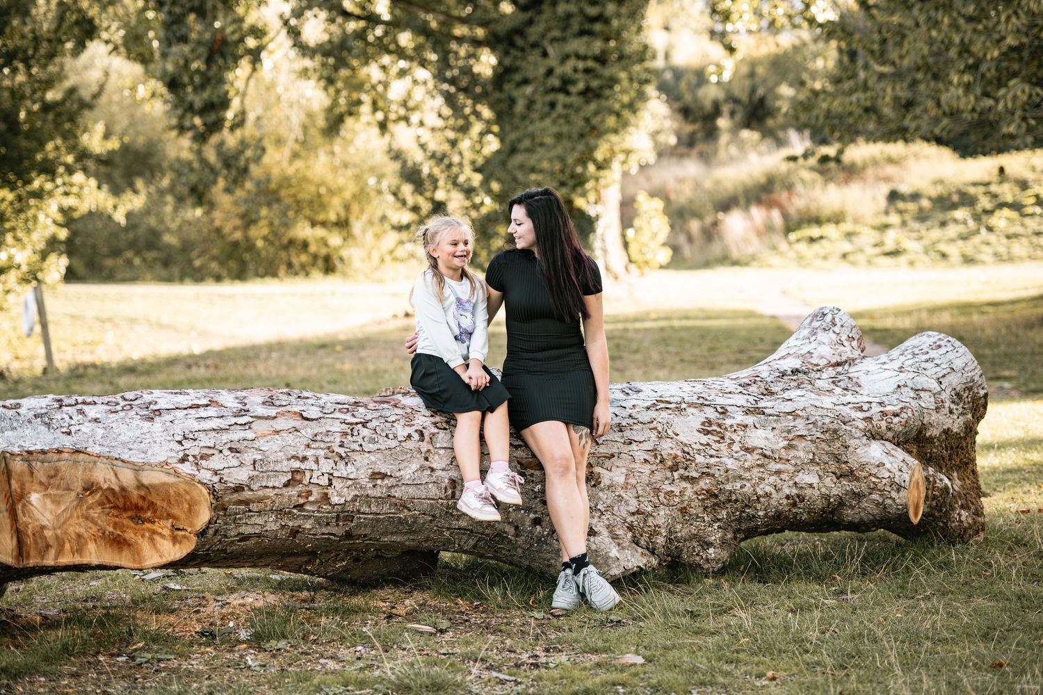Two people walking along a fallen tree trunk in a sunlit park setting.