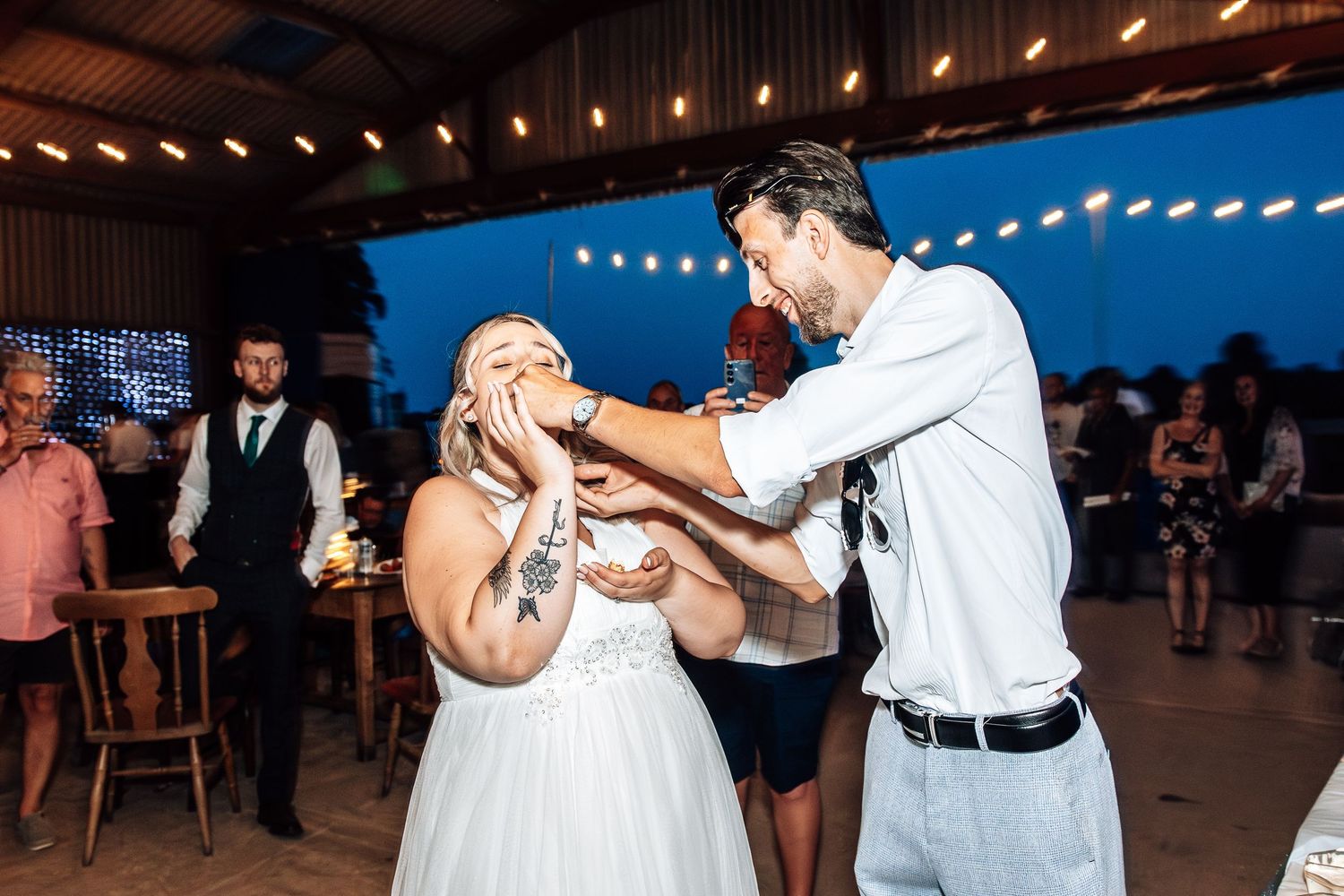 Couple dancing and laughing together during an evening reception under string lights at an outdoor venue.