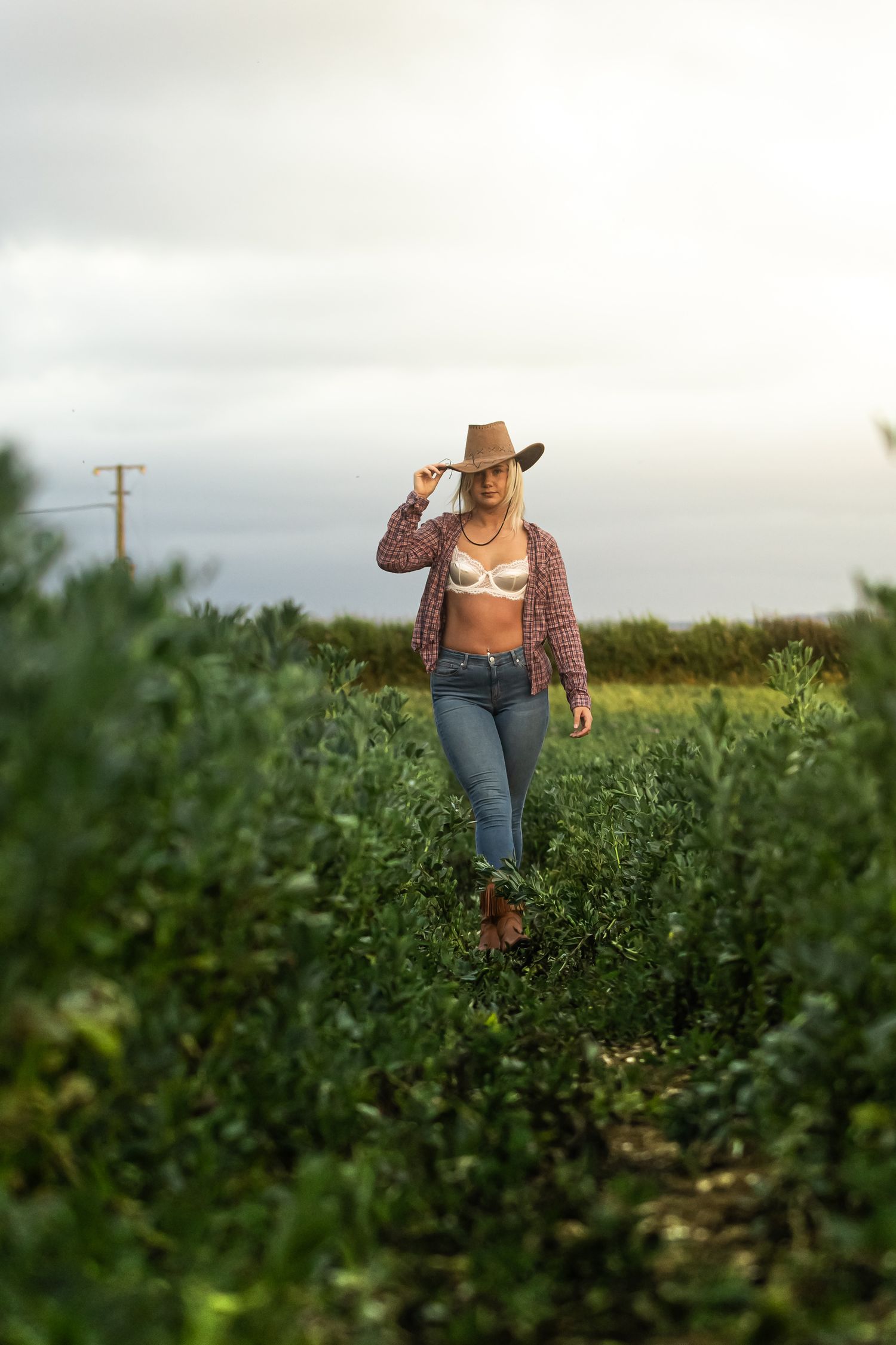 A person in a brown hat and cardigan walks through a lush green field on an overcast day.