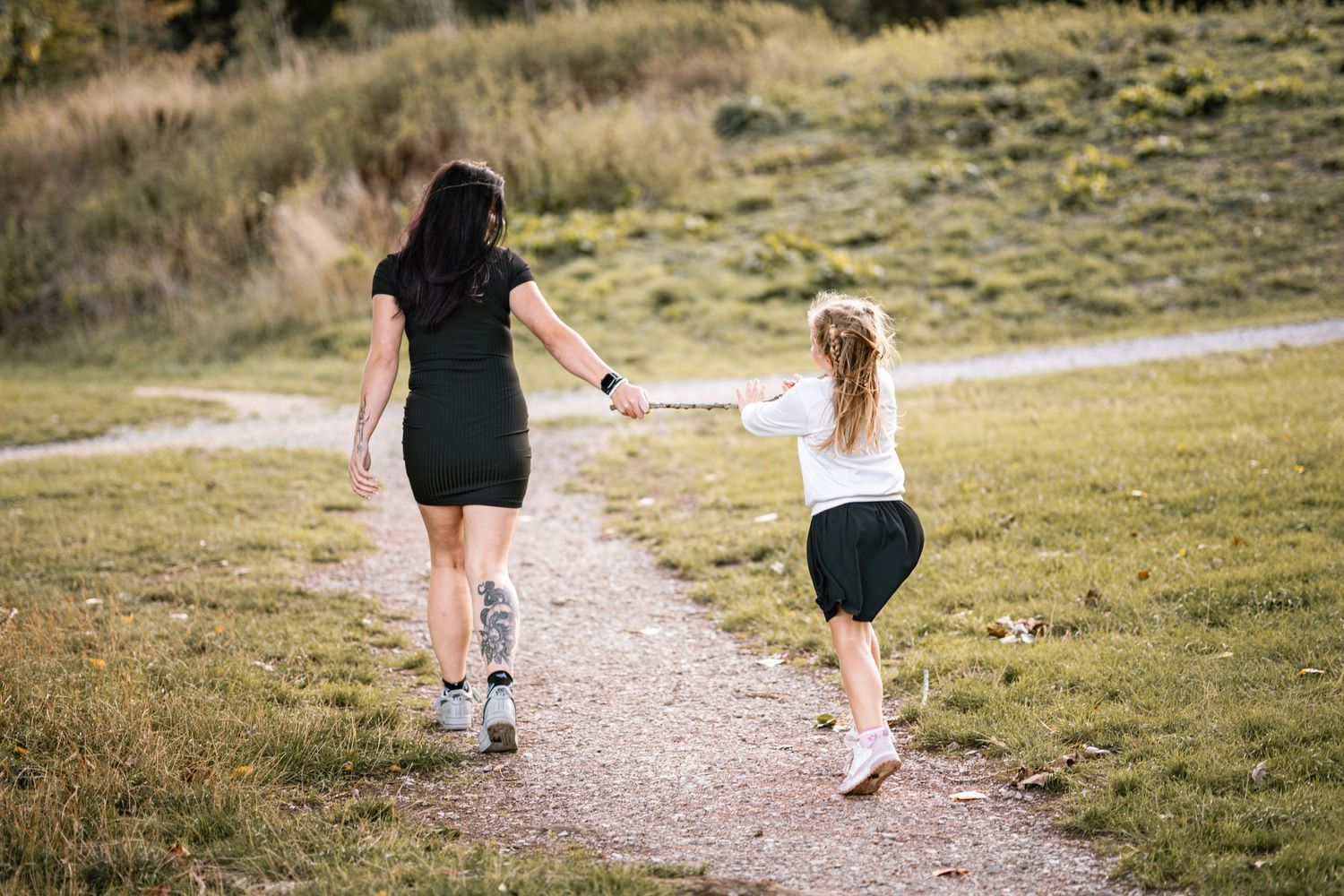 Two people walk together along a winding gravel path through a grassy hillside on a sunny afternoon.