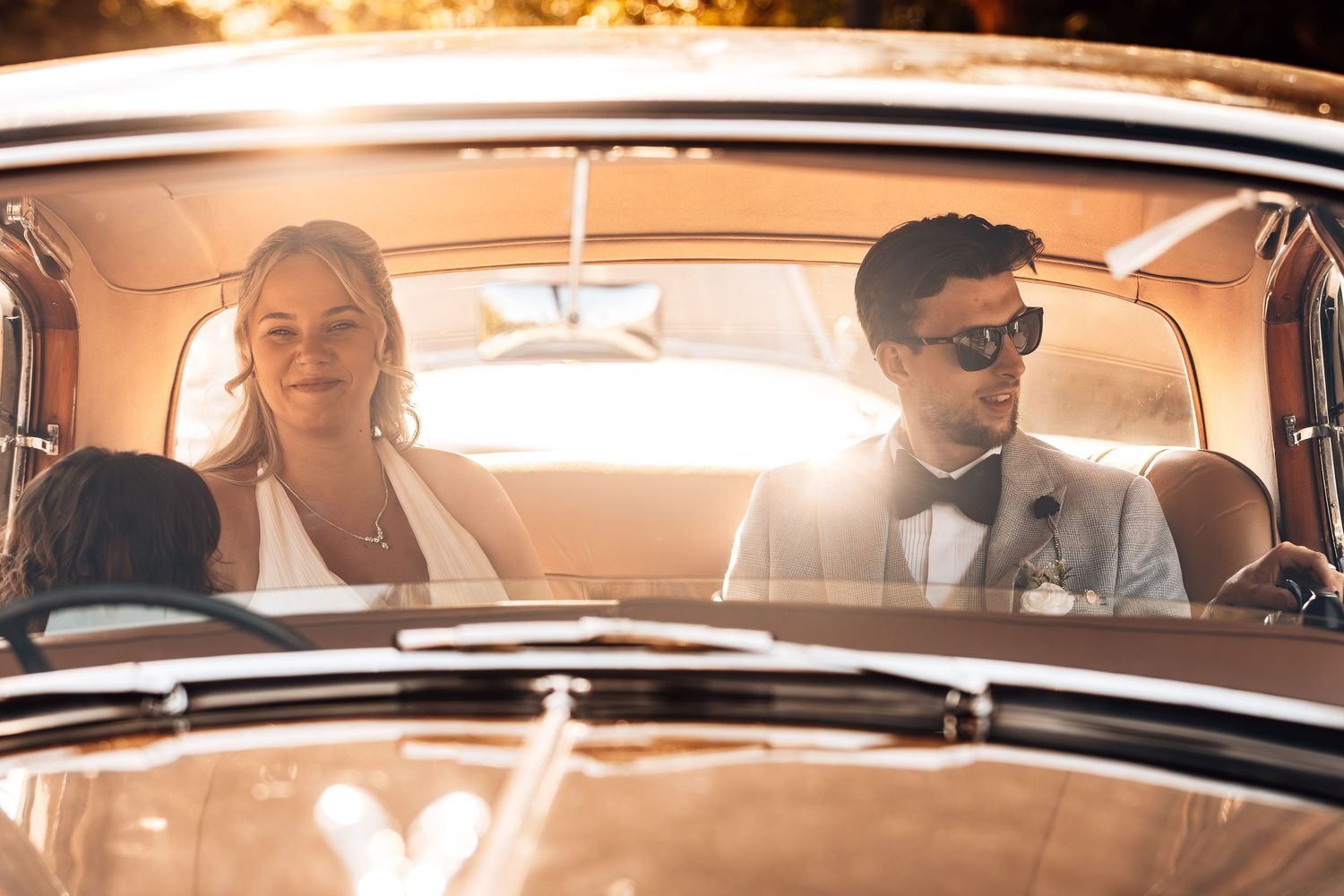 A group of people sit in a classic car with warm lighting filtering through the windows during a vintage-style photoshoot.