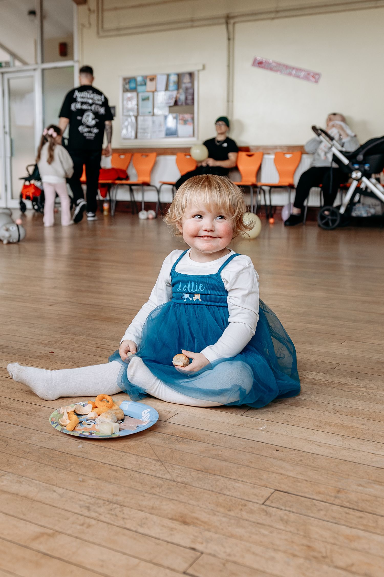 A small child in a blue dress and white shirt sits on a wooden floor in a dance studio.