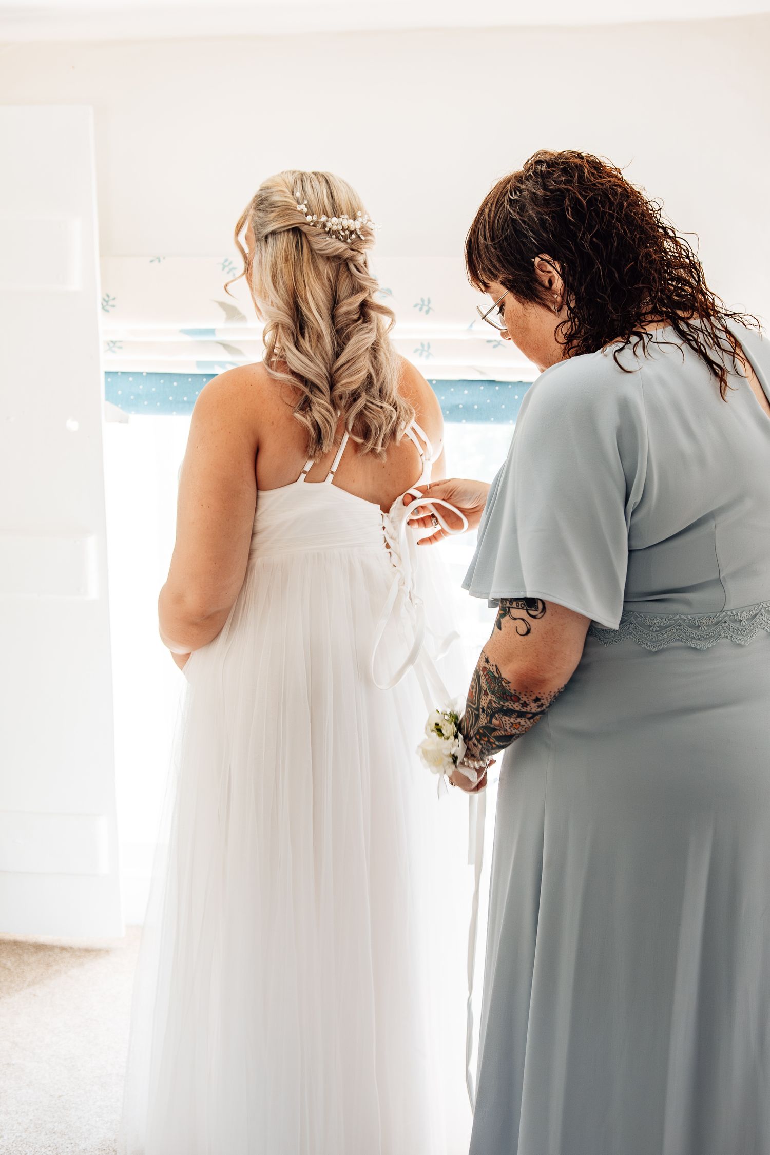 A bridesmaid in sage green helps adjust a white wedding dress with intricate braided hairstyle details.