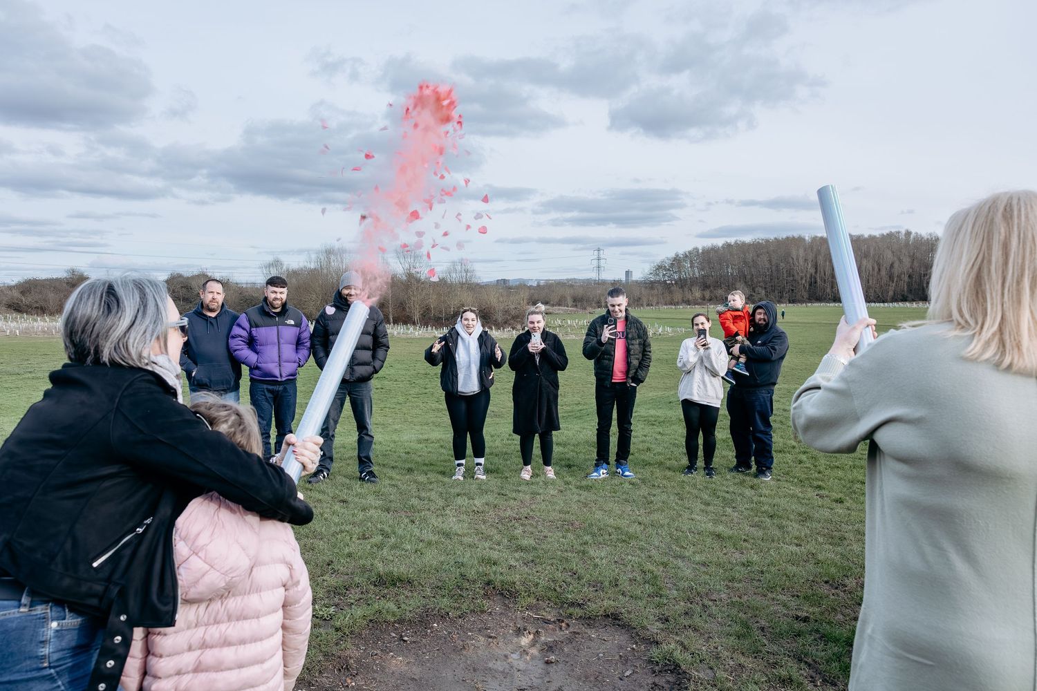 A group gathers outdoors for a gender reveal celebration with pink smoke floating in the cloudy sky.