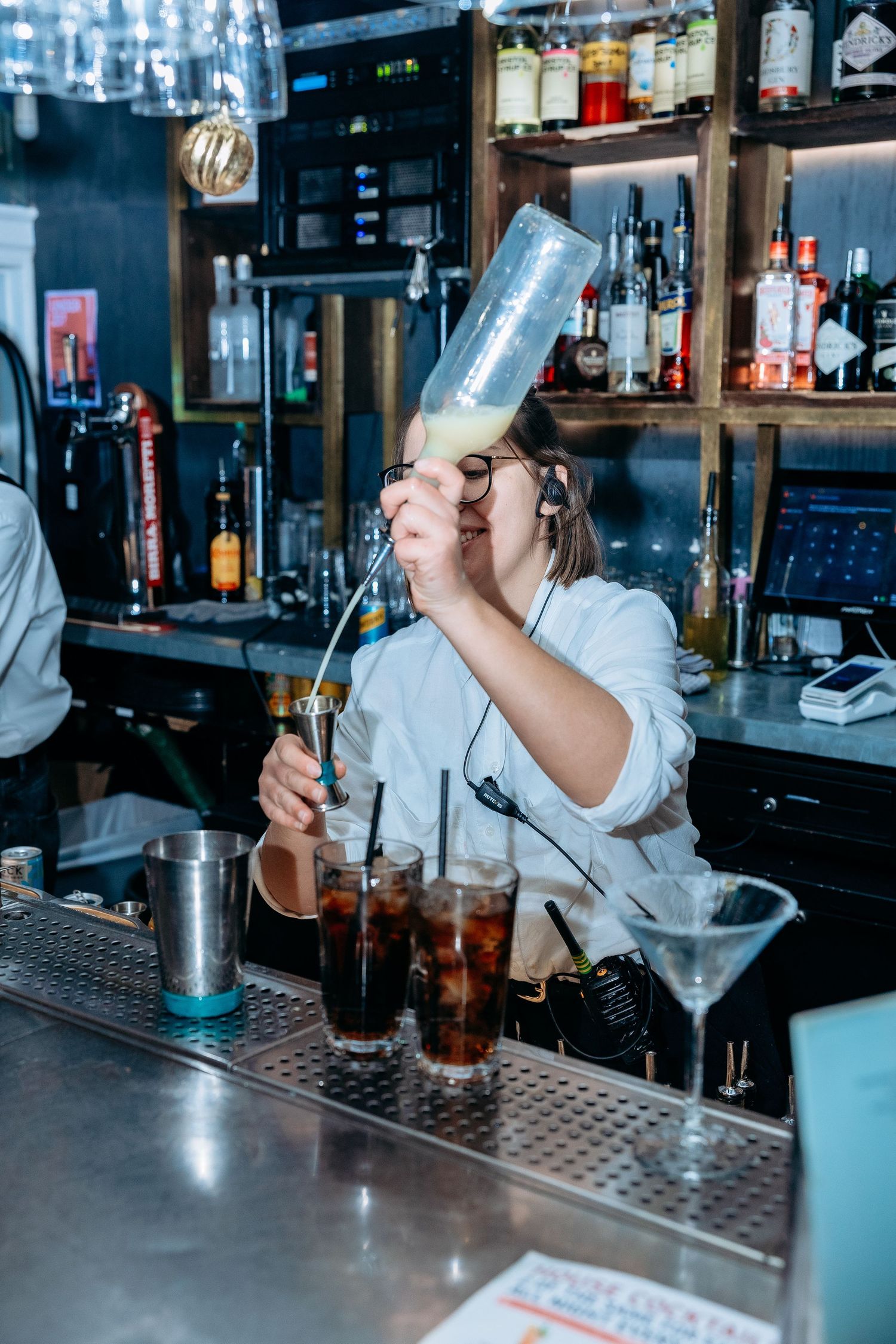 Bartender shaking cocktails at a modern bar counter with bottles and equipment in the background.