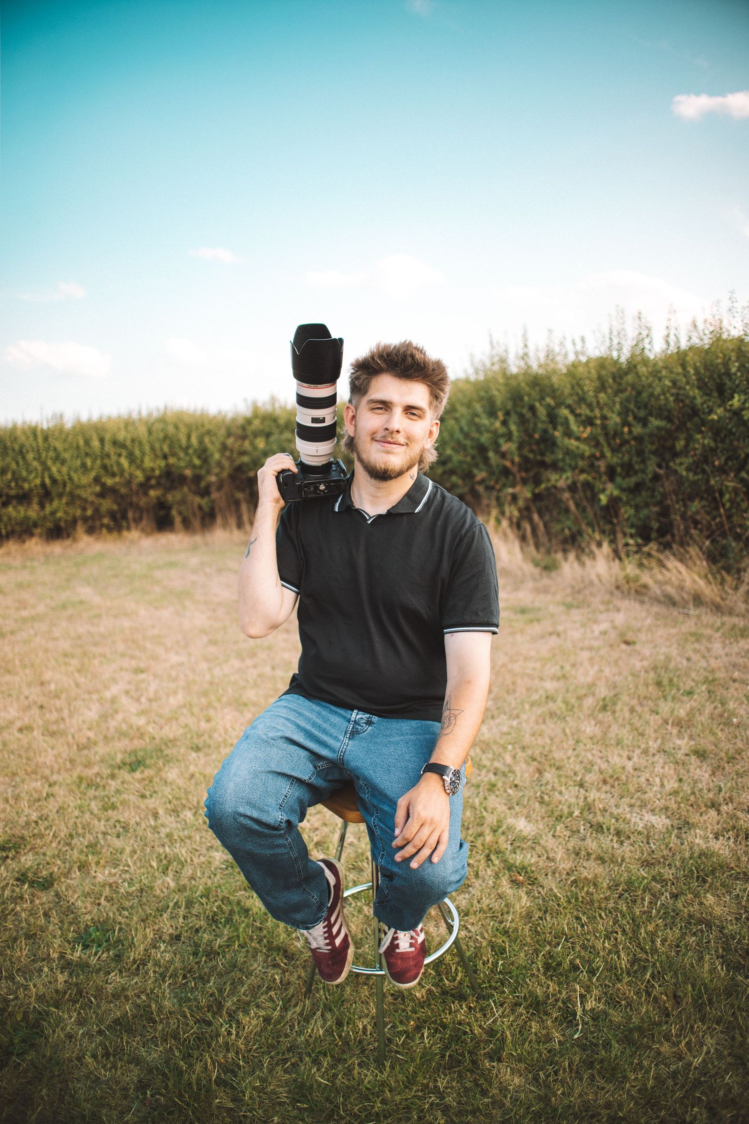 Photographer crouching in grassy field holding camera against blue sky backdrop.