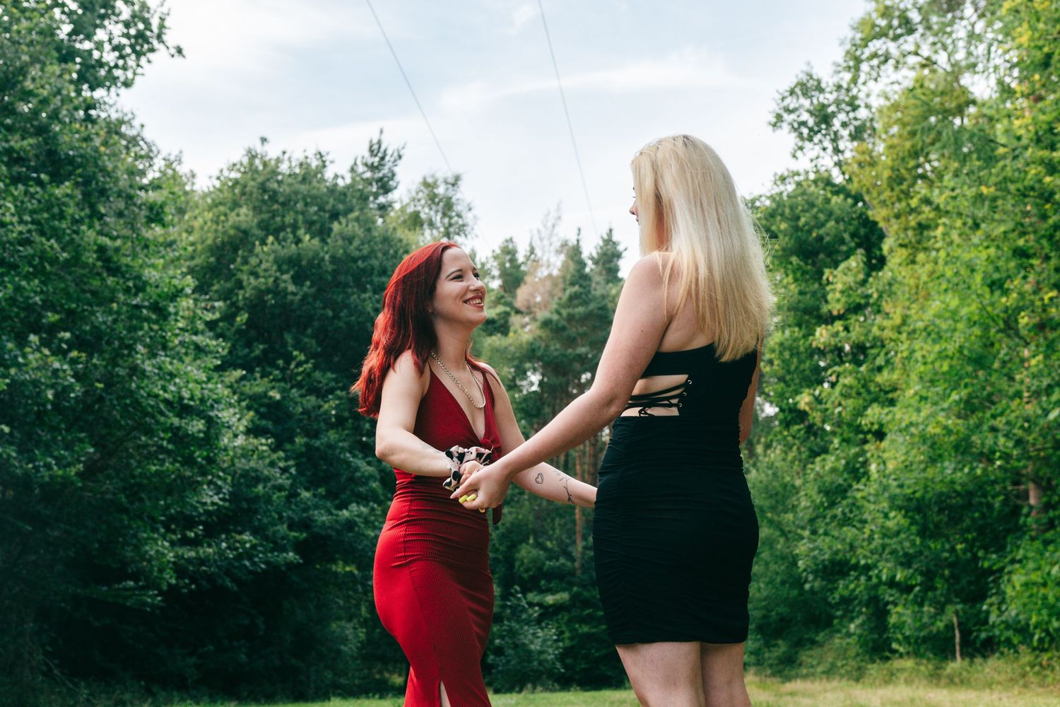 Two figures in red and black dresses dance together in a lush green outdoor setting during summer.