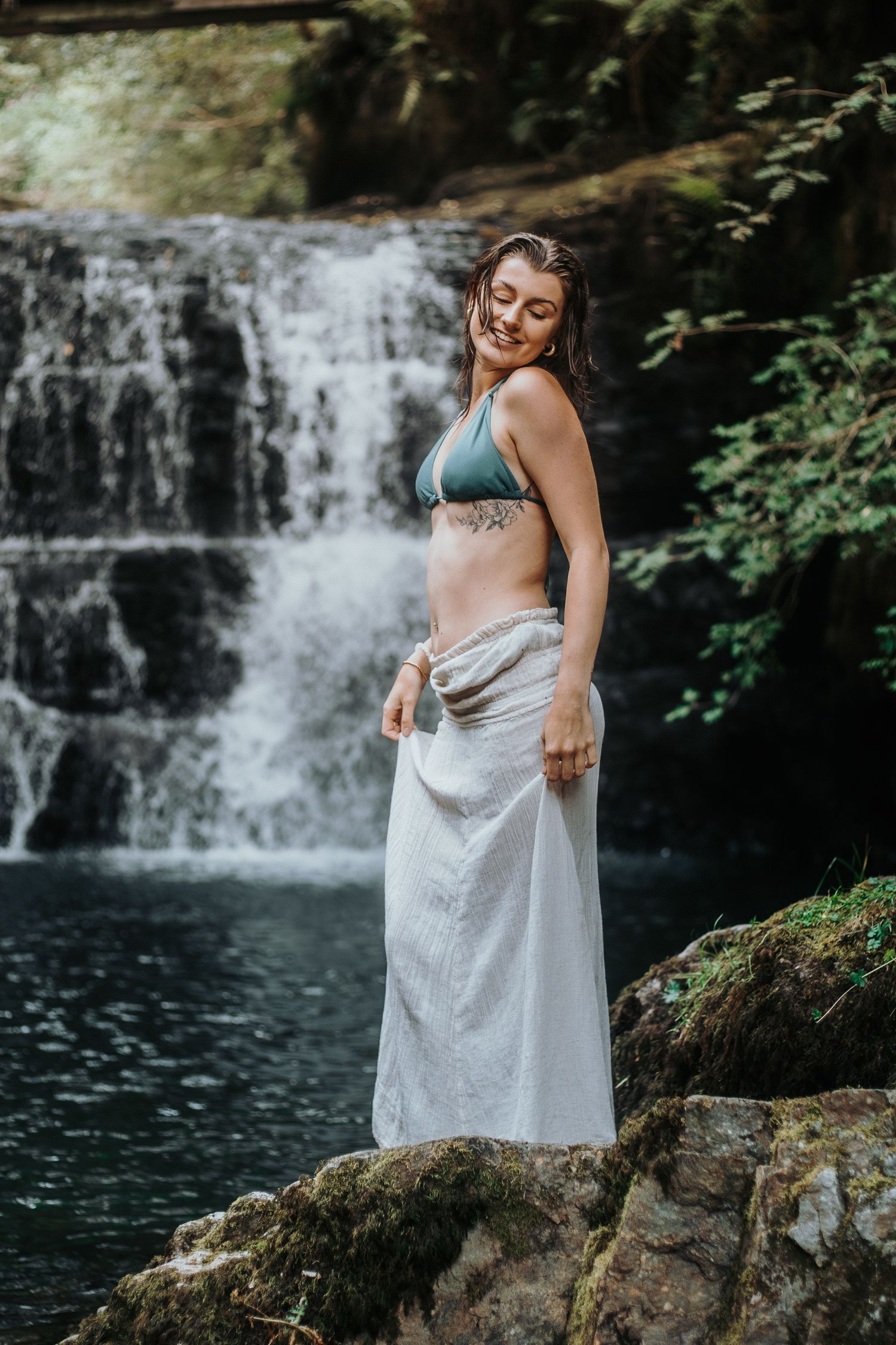 Maternity photoshoot by a cascading waterfall with a person in a white dress posing on mossy rocks near dark water.