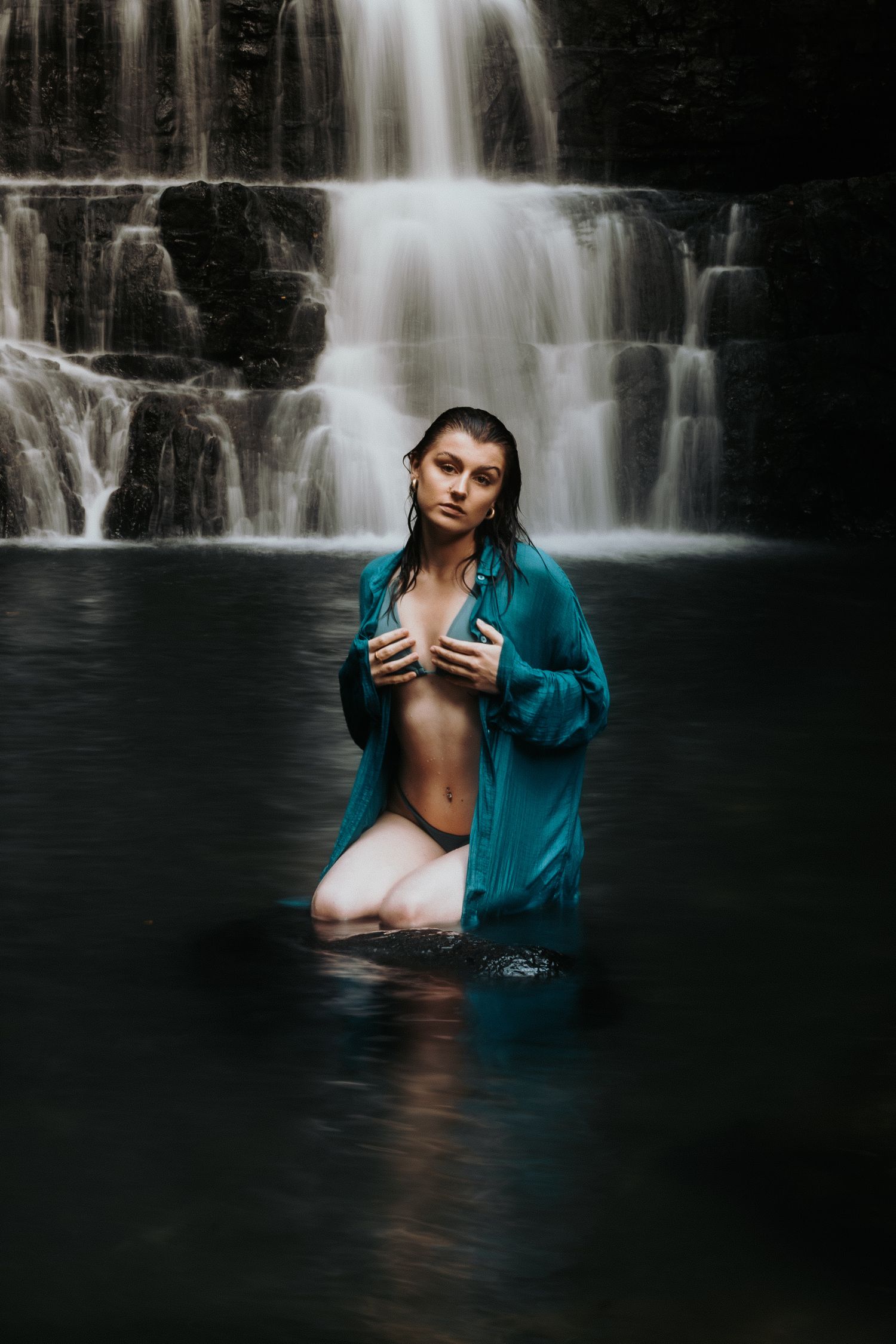 Artistic portrait in dark water against cascading waterfall backdrop with teal shirt and moody lighting.