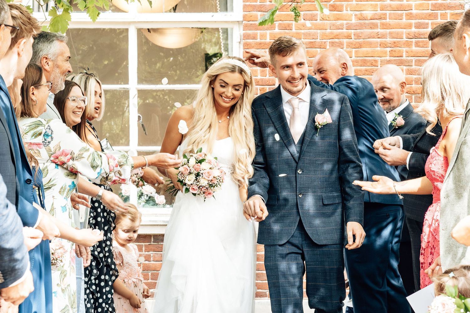 Wedding guests celebrate outside brick building during daytime reception.