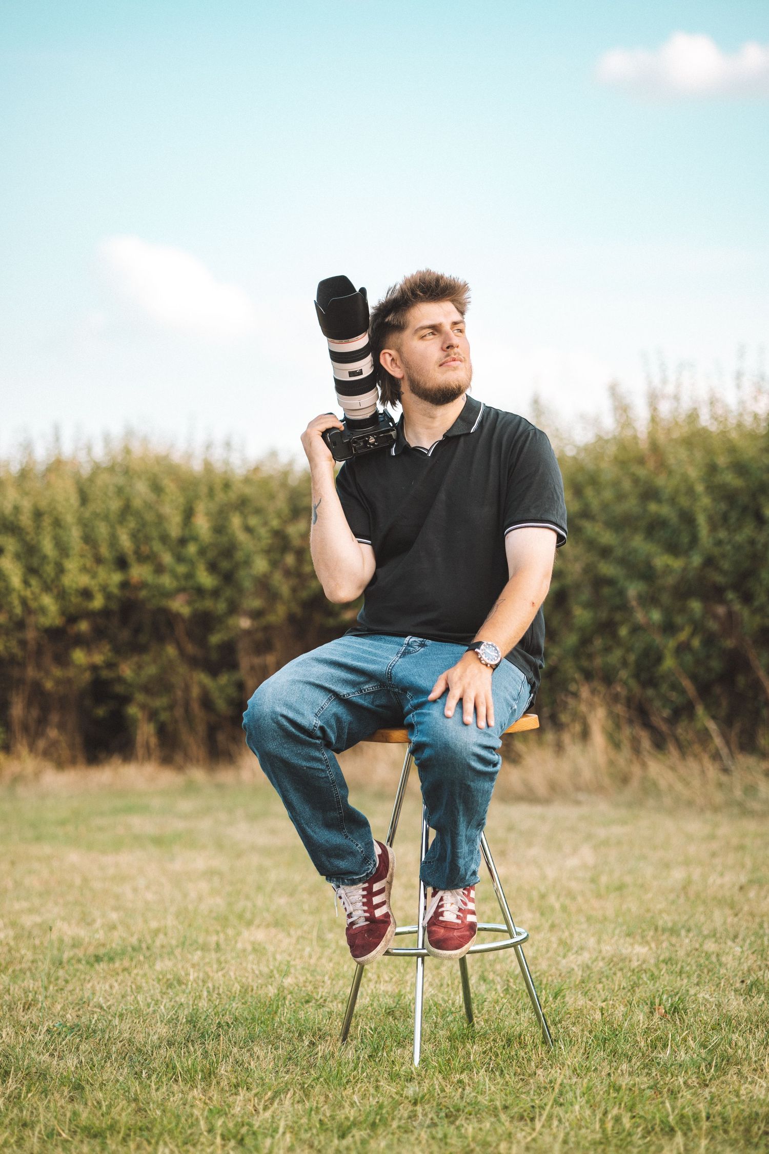 Photographer sitting on chair in grassy field holding professional camera equipment.
