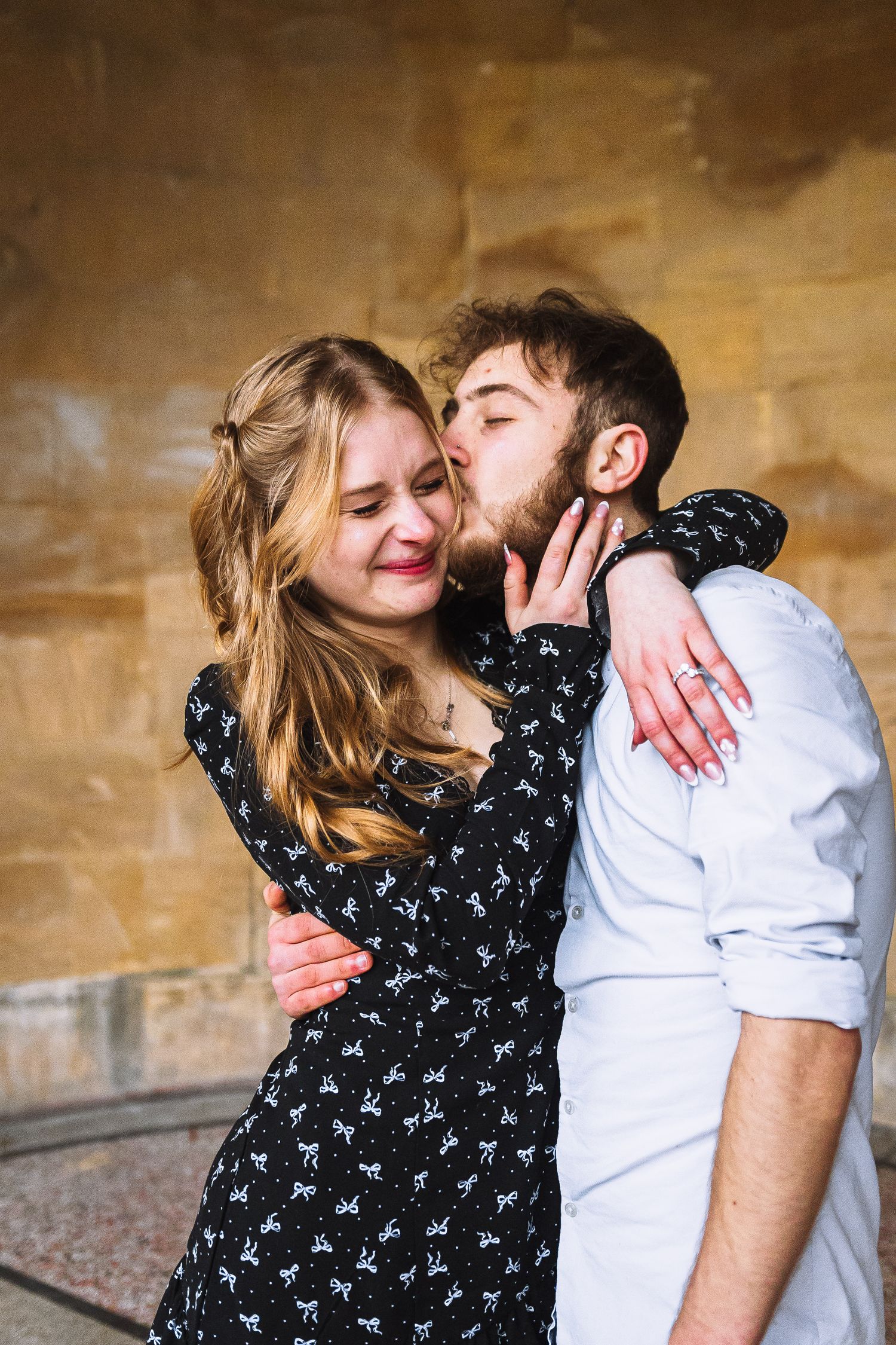 A couple shares an intimate moment against a golden wall, with her in a patterned black dress and him in a white shirt.