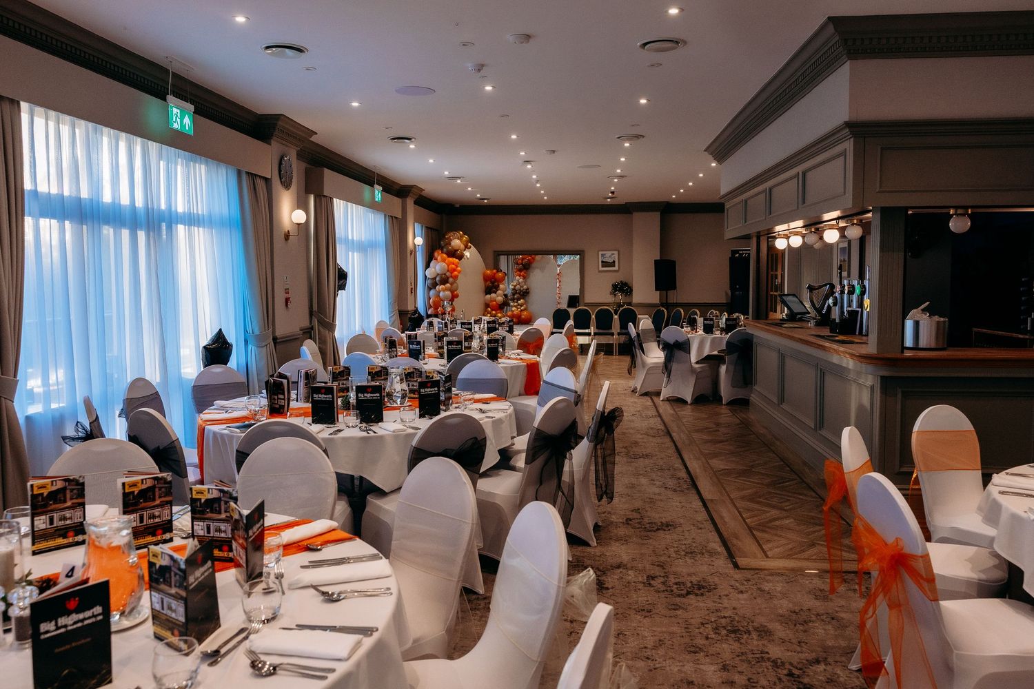 Modern banquet hall with white chairs and brown carpeting features elegant table settings and panoramic windows.