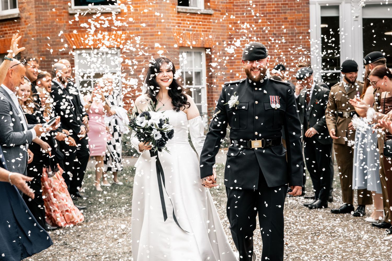 Wedding guests shower newlyweds with white confetti outside brick building.