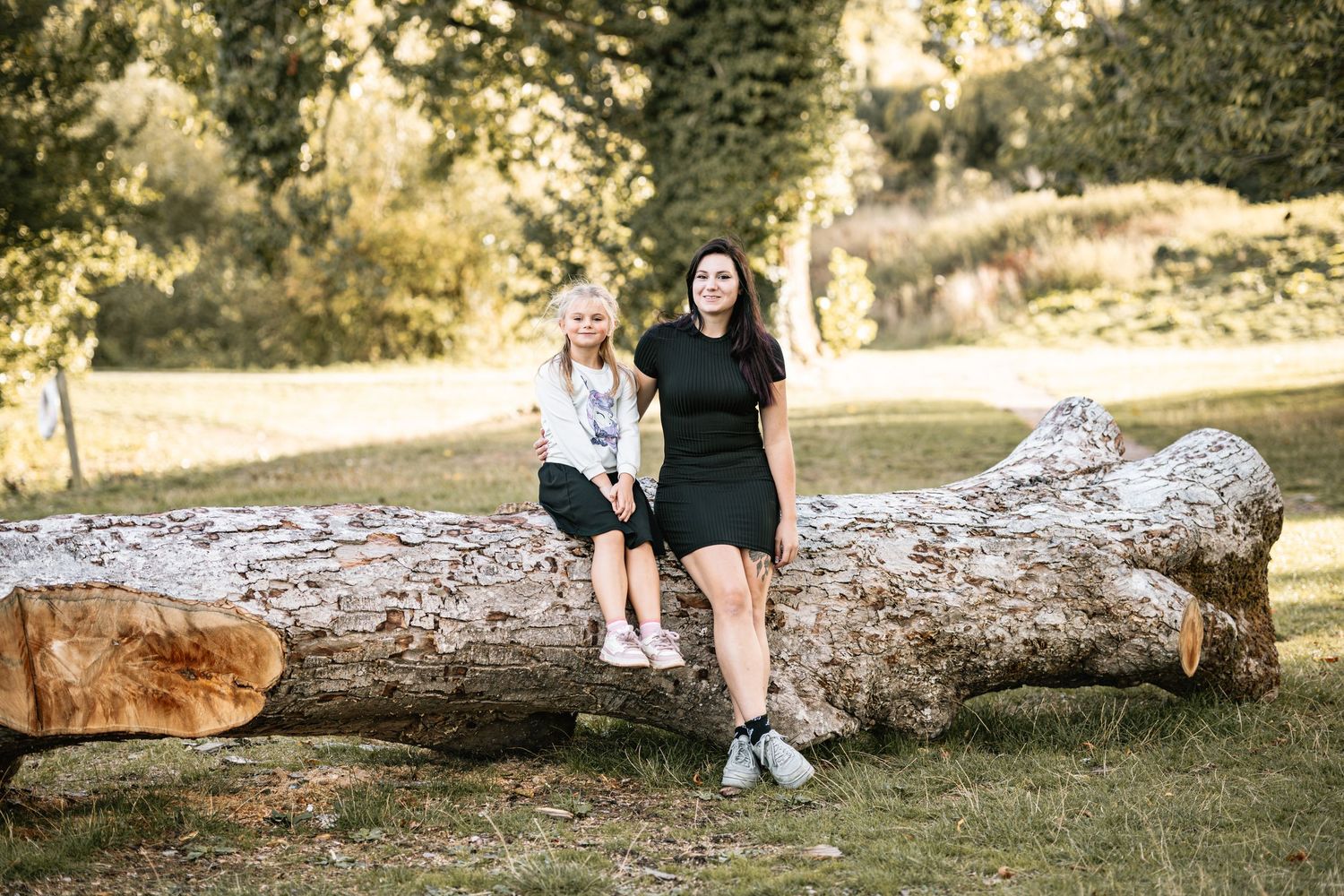 Two people sit together on a large fallen log in a sunny park setting with trees in the background.