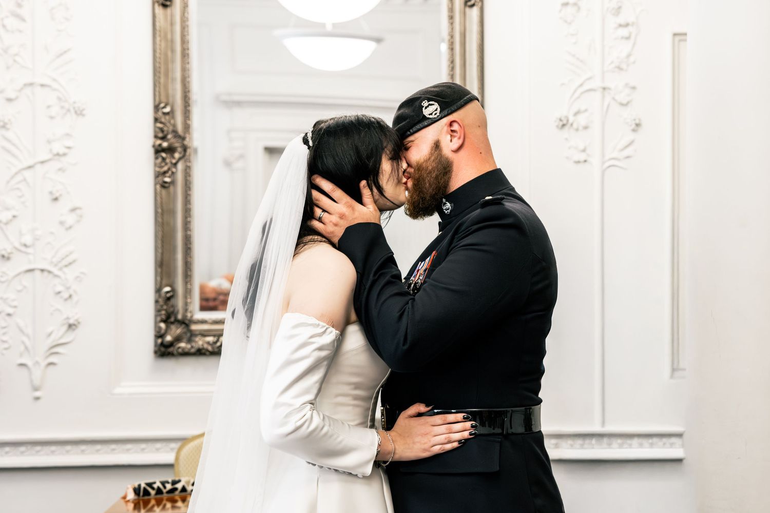 A romantic wedding couple shares a passionate kiss against an elegant white wall with ornate molding details.