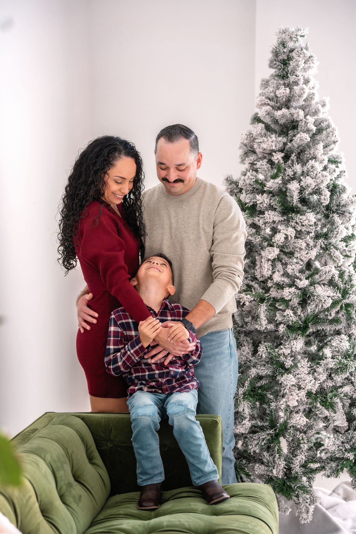 A couple shares intimate moments on a green velvet couch with snow-flocked Christmas trees in the background.