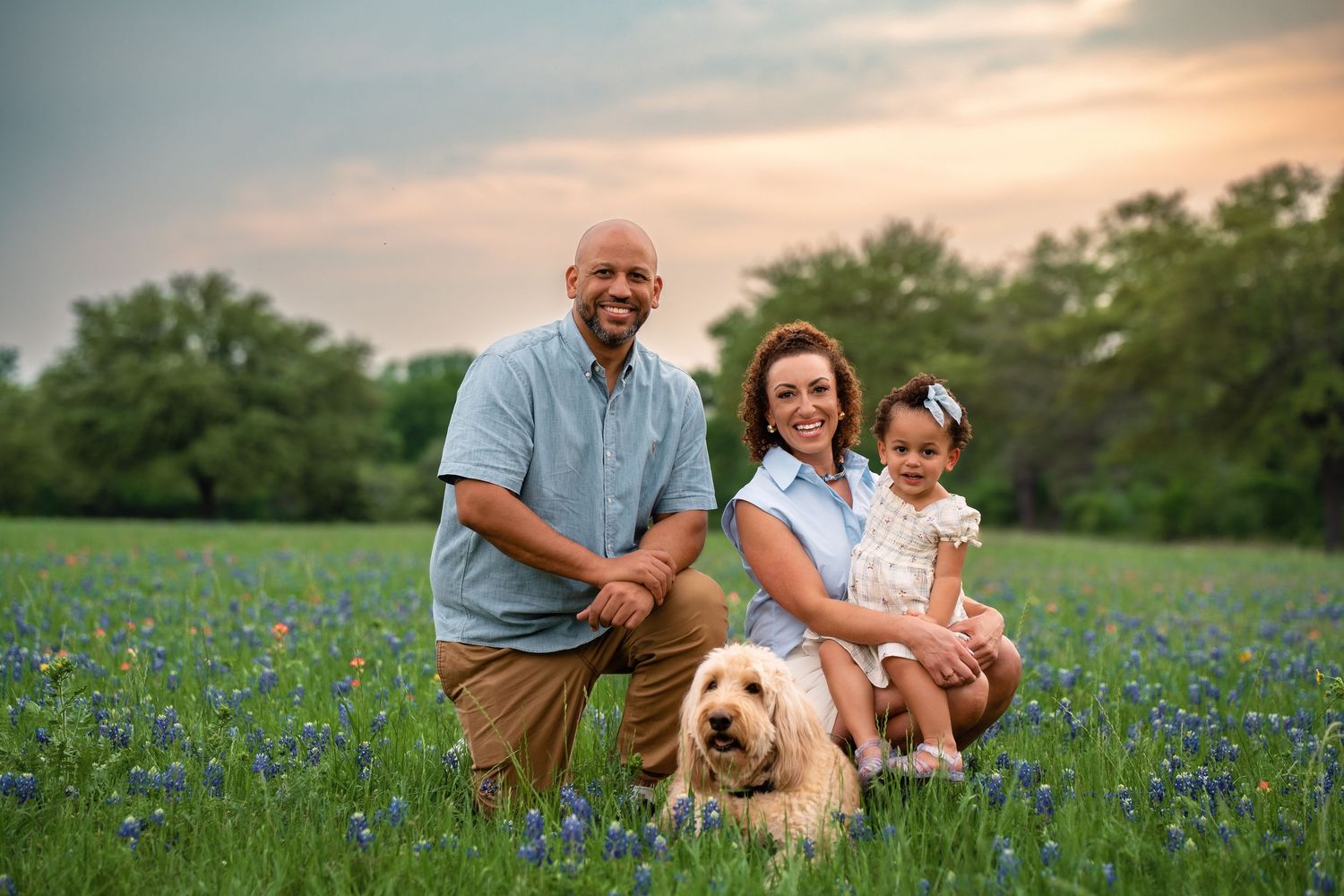 Family and their golden retriever dog sitting together in a blooming field of bluebonnets at sunset.