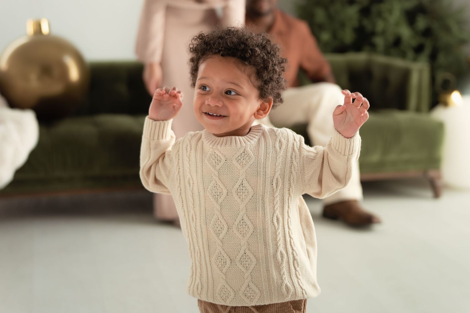 Happy toddler in cream cable-knit sweater smiling with raised hands in a cozy living room.