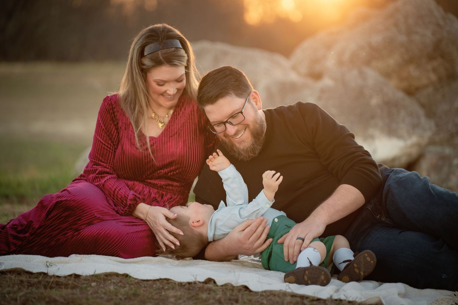 A young family sits on a blanket in Buda, TX at sunset, parents smiling at their toddler lying between them.
