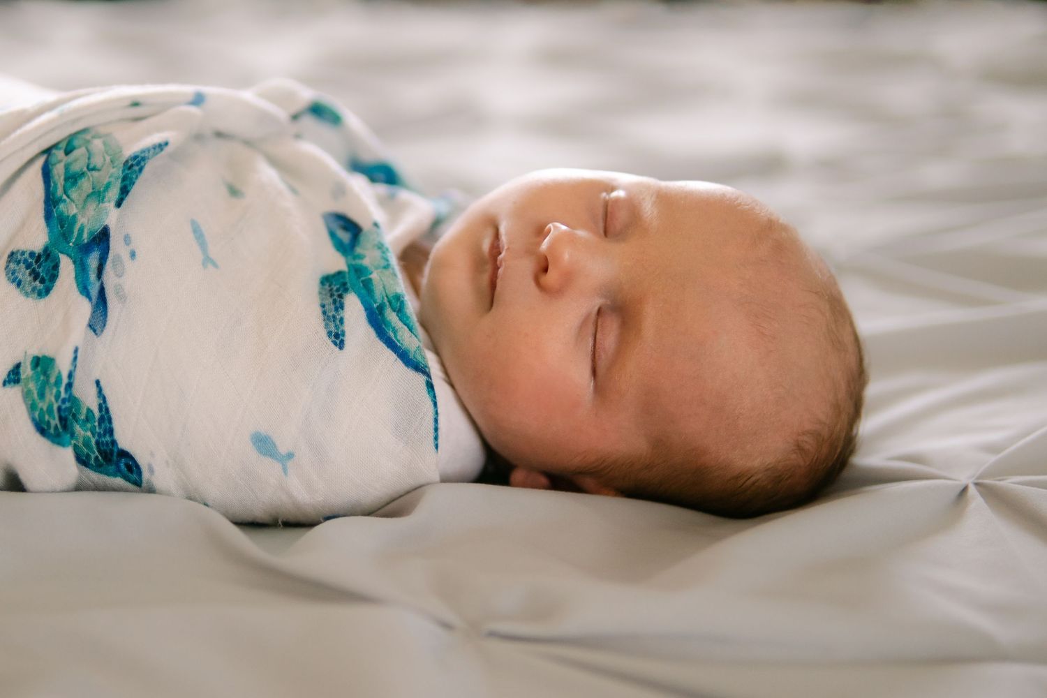Newborn baby sleeping peacefully swaddled in a white blanket with blue turtle print on a soft gray surface.