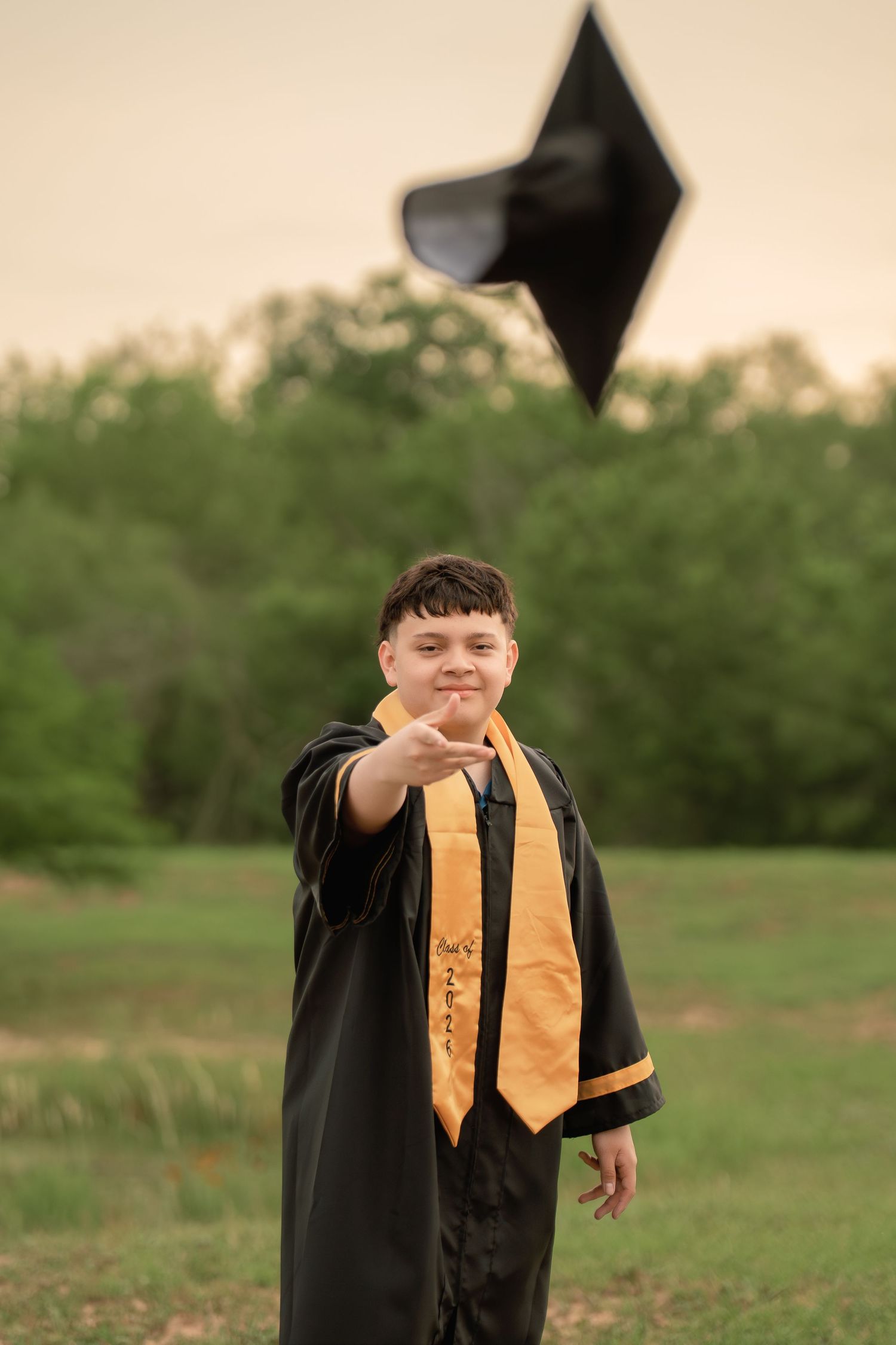 A smiling graduate in black gown and gold stole tosses cap in air outdoors.