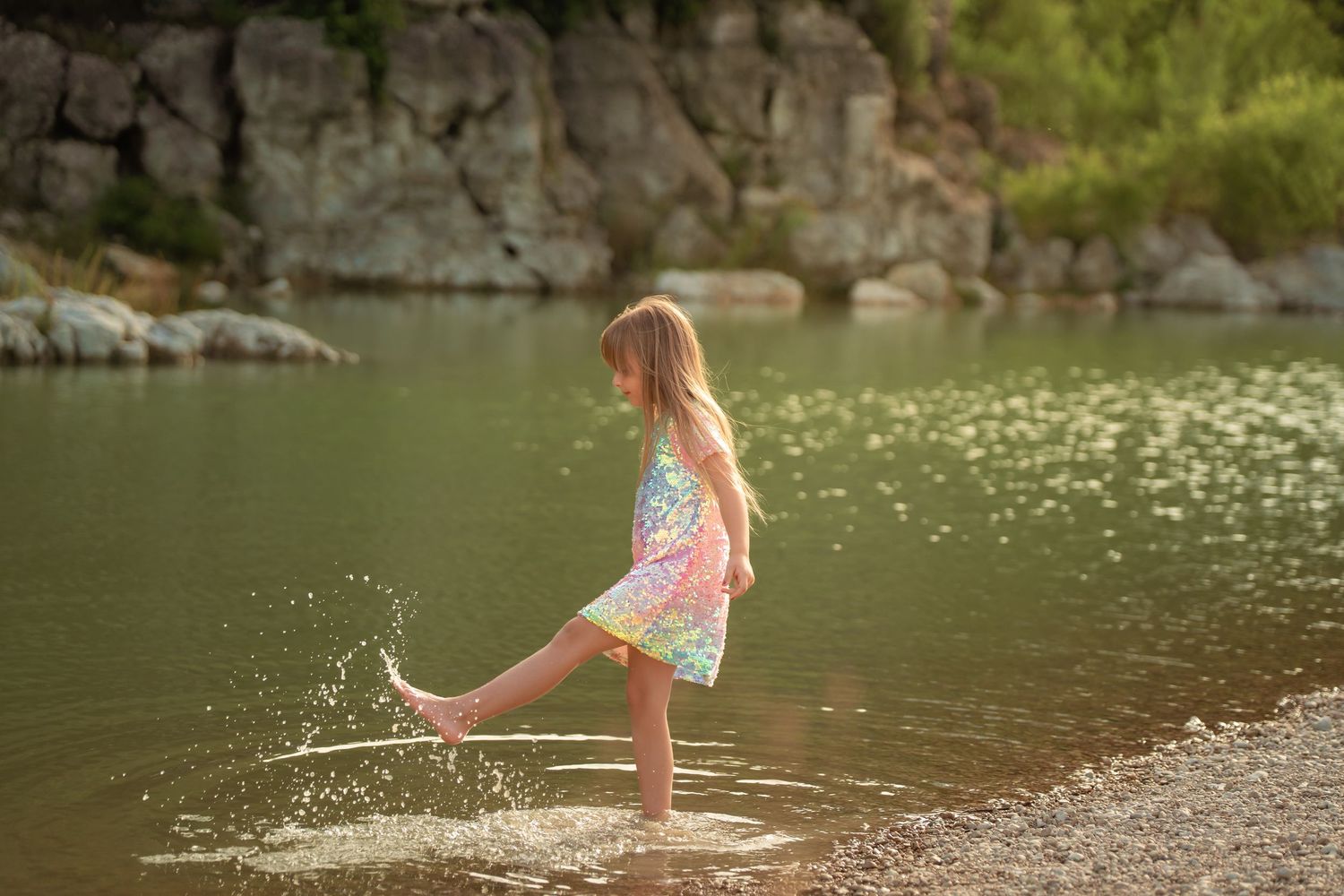Young girl in colorful dress splashing water with her foot at a rocky lake shore in golden sunlight.