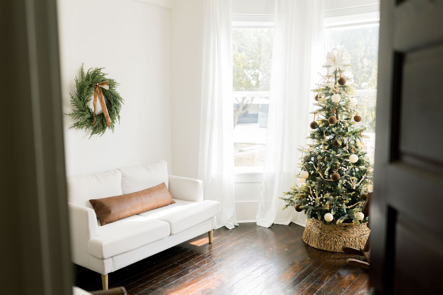 Bright living room with white sofa, Christmas tree, and festive wreath on wall with hardwood floors.