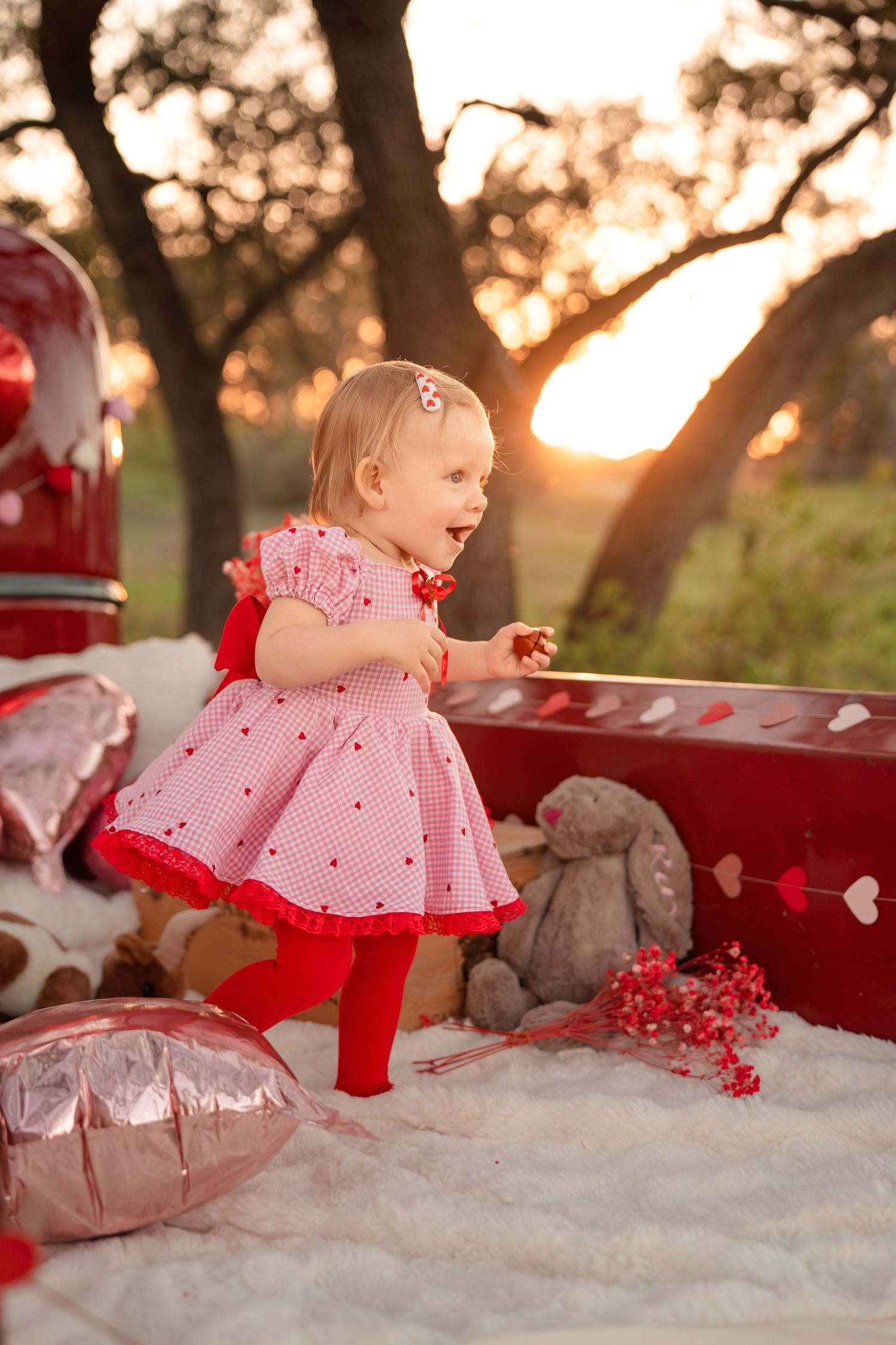Toddler girl in a pink gingham dress and red tights laughs on a white blanket at a Valentine's Day sunset photo shoot with heart balloons.