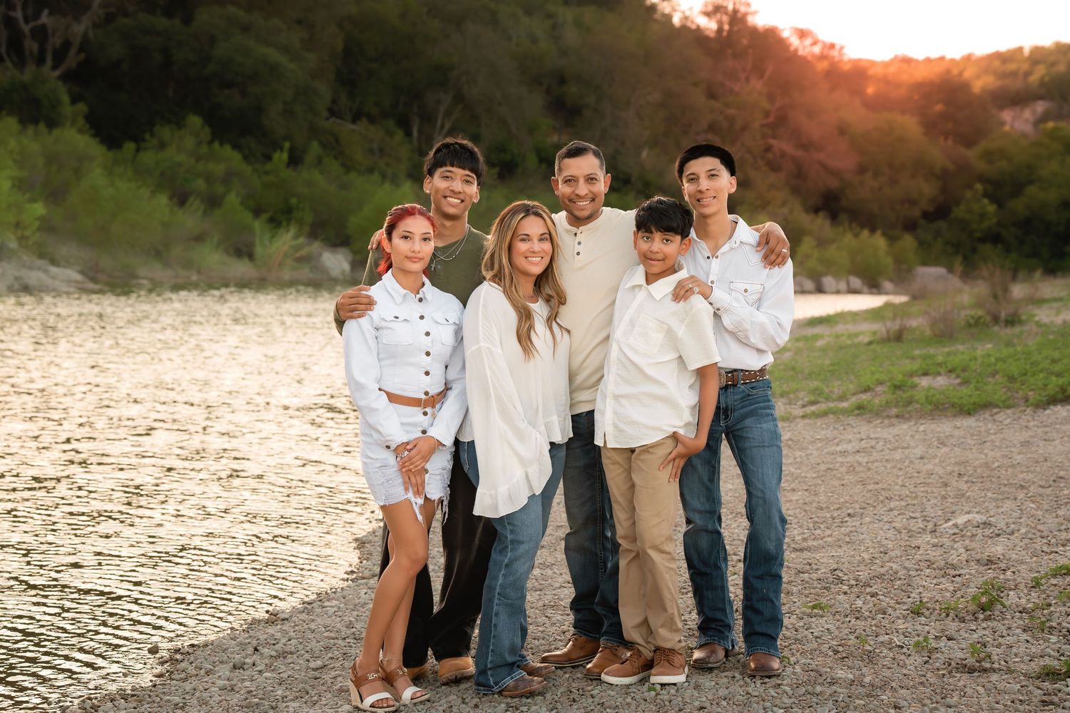 Family of six posing together on a rocky riverbank at golden hour, wearing coordinated white tops and jeans.