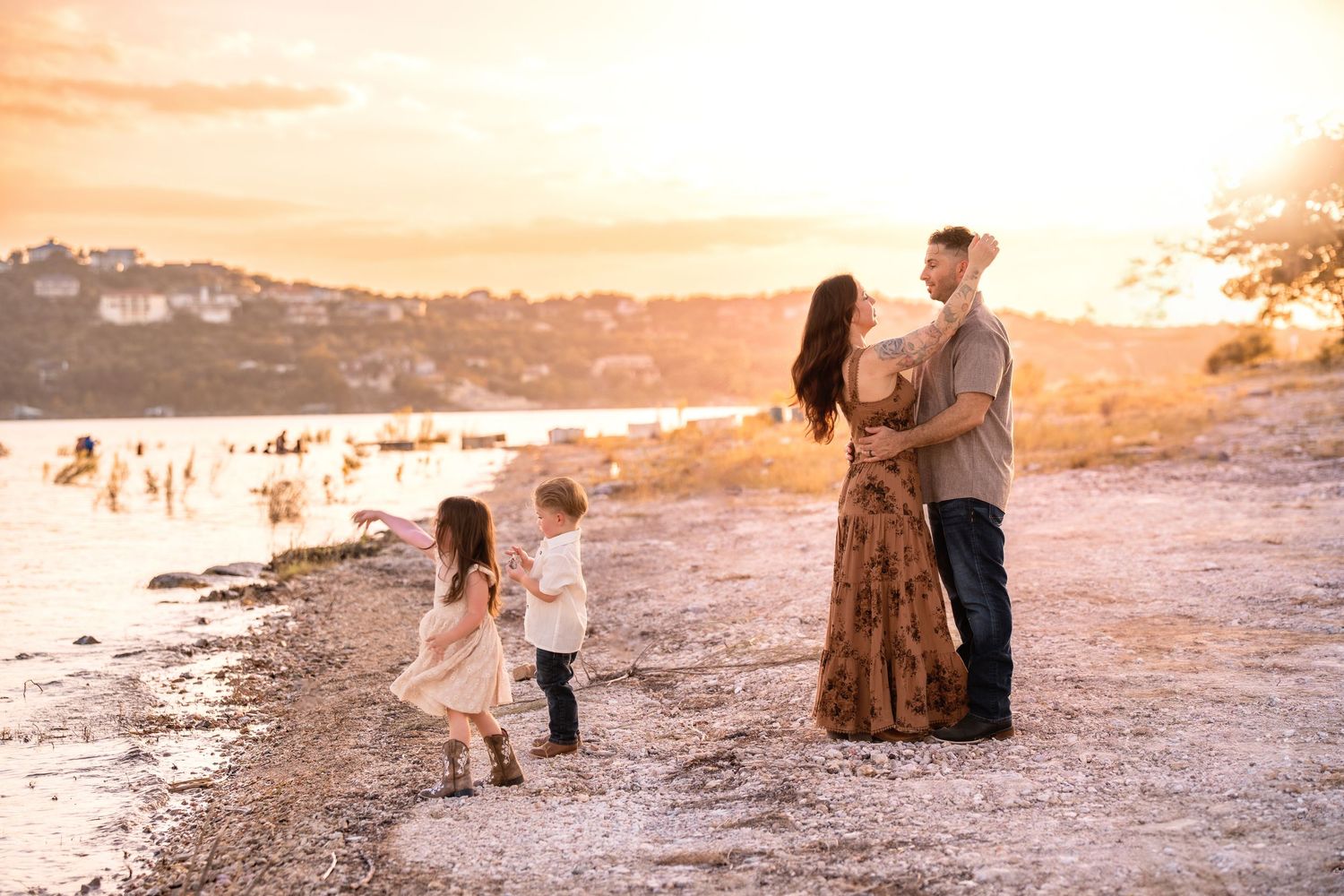 Family of four enjoying a golden sunset on a rocky beach, parents embracing while two young children play nearby.