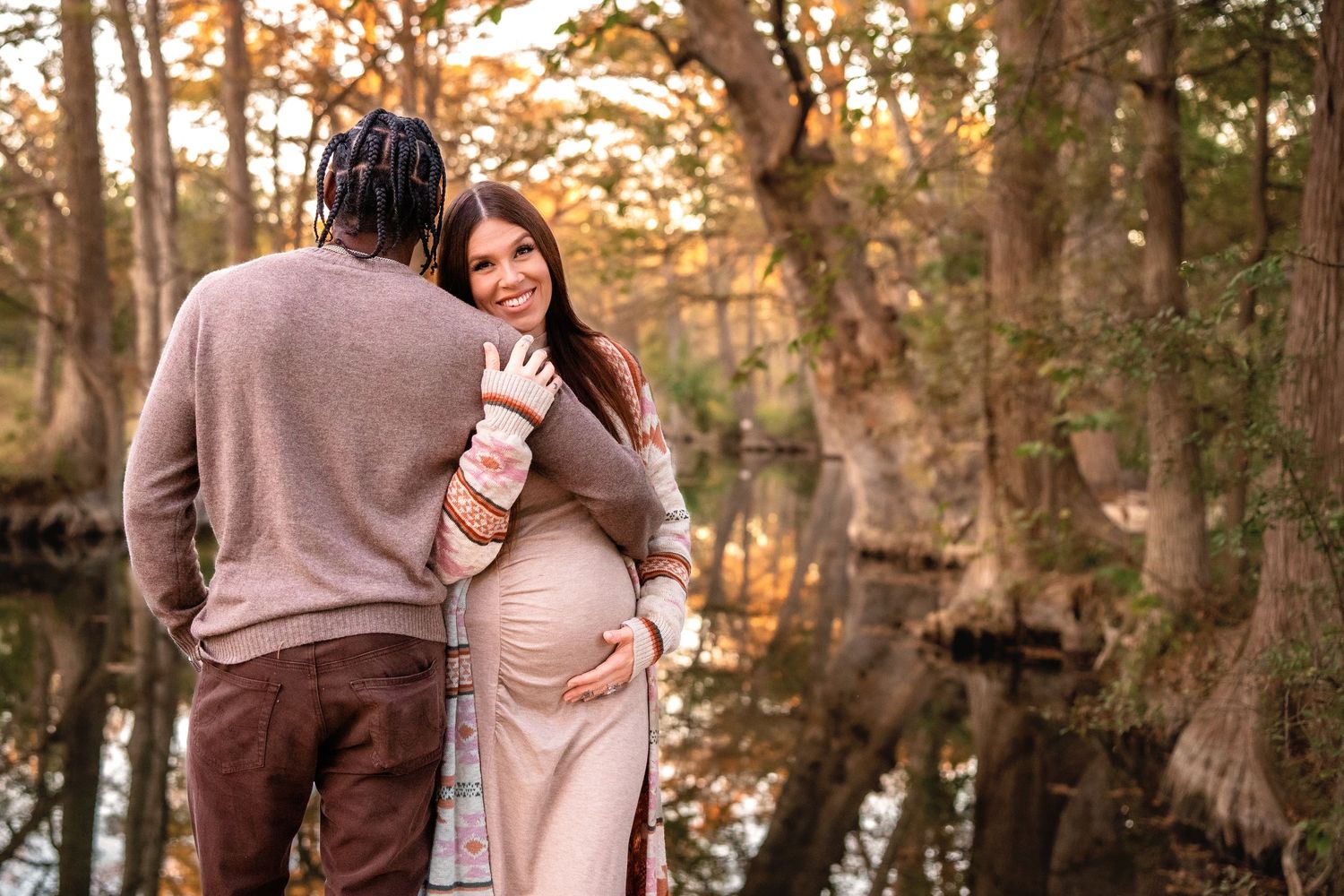 Pregnant couple embracing outdoors near a reflective pond surrounded by autumn trees in warm golden light.