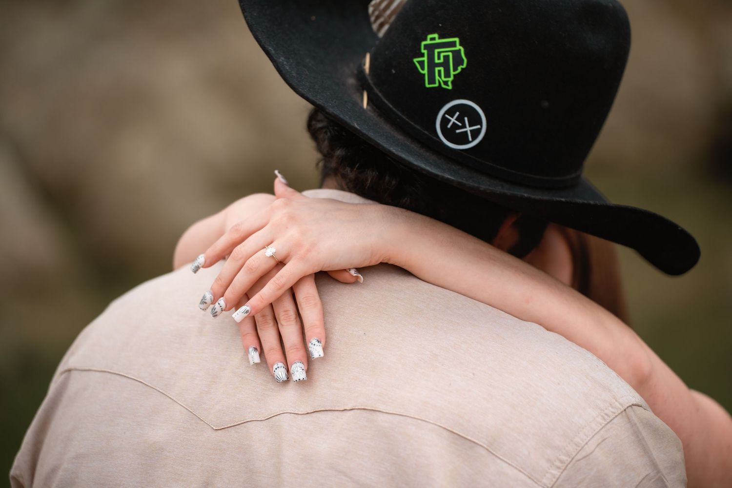 A woman with manicured nails rests her hand on a man's shoulder, wearing a black cowboy hat with green logo.