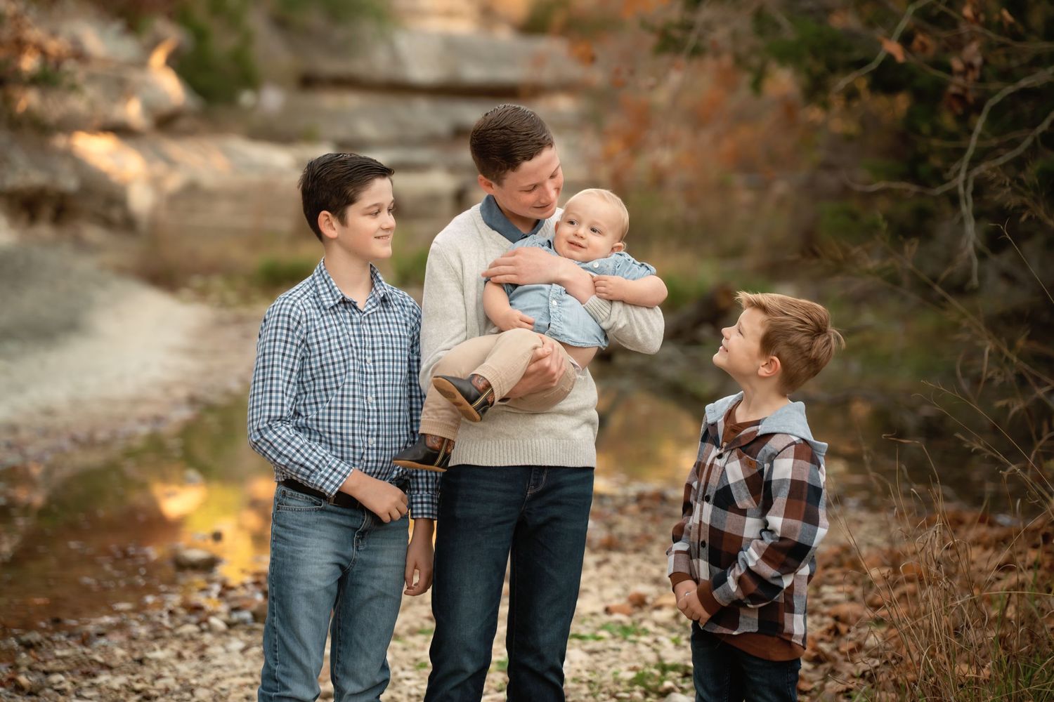 A father holds a baby outdoors in fall while two boys stand nearby smiling in a warm family portrait.