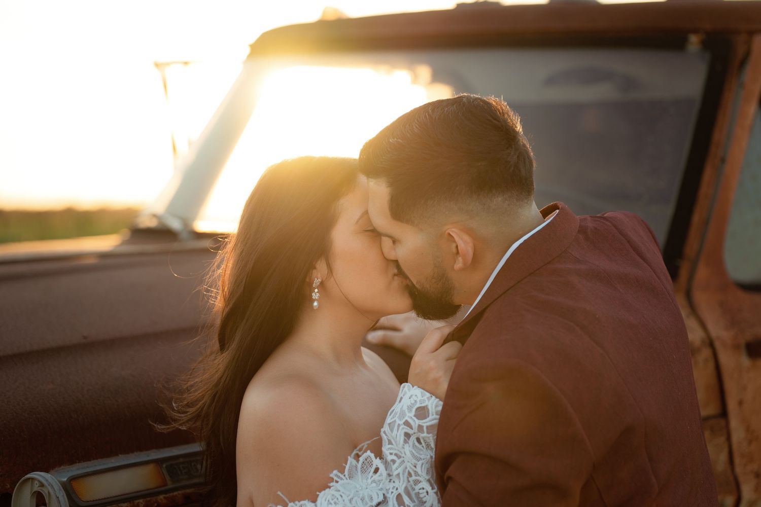 A romantic couple shares an intimate kiss in a sun-lit car during their wedding day celebration.
