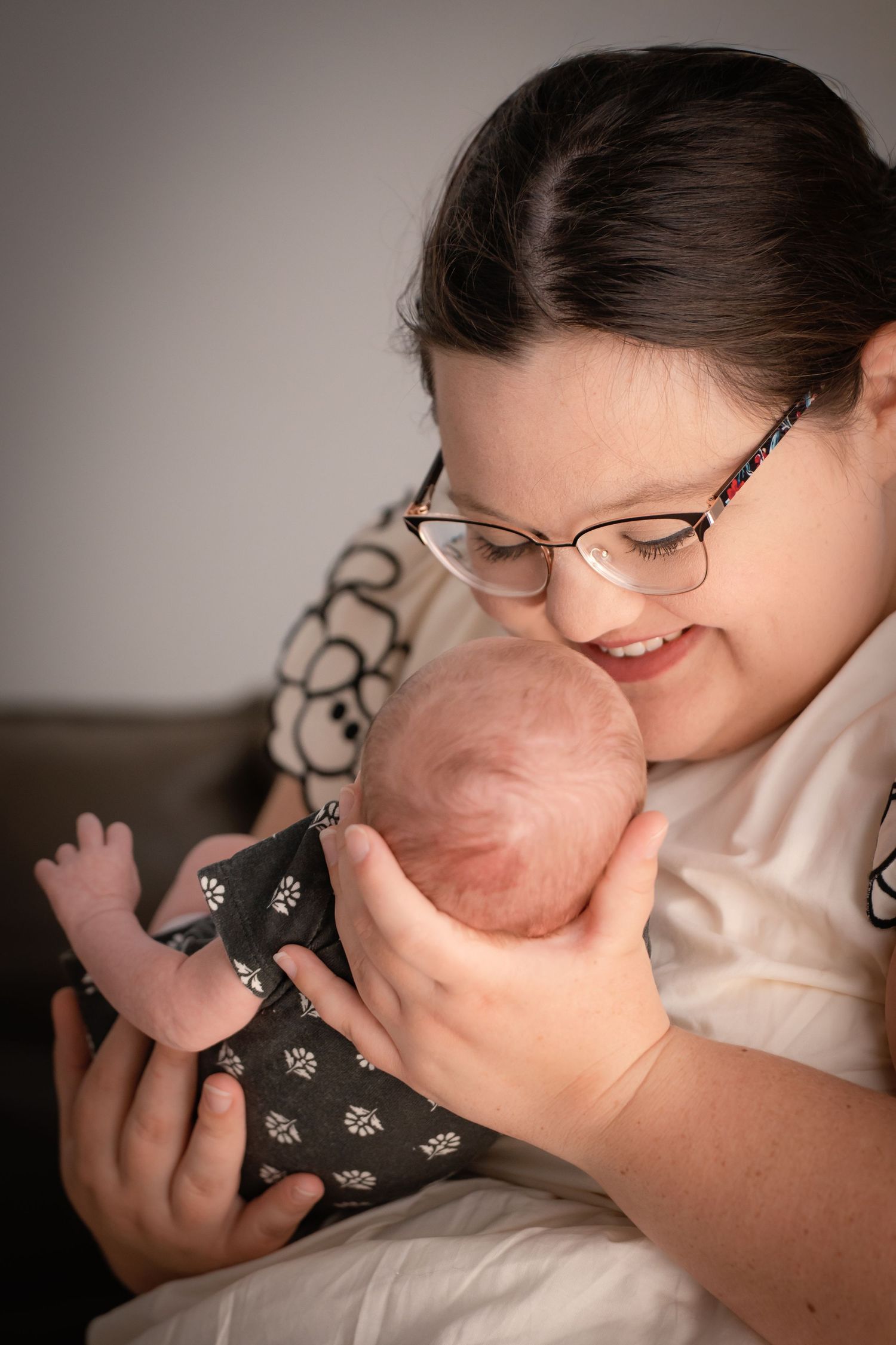 Smiling woman with glasses tenderly holds a sleeping newborn baby wrapped in a dark floral blanket.