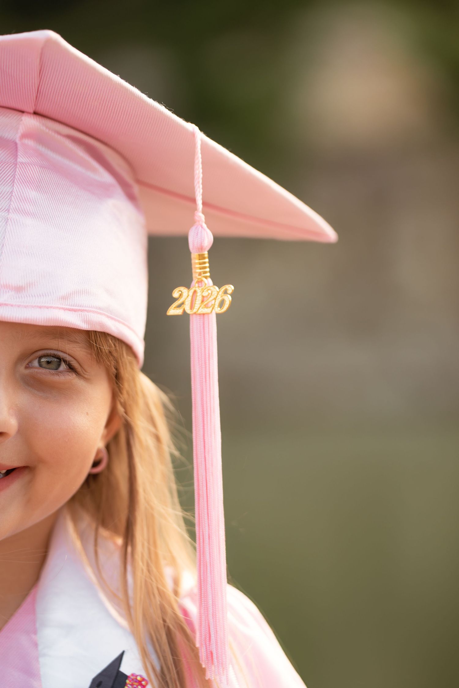 Young girl wearing pink graduation cap with 2026 tassel, smiling in her kindergarten graduation ceremony.