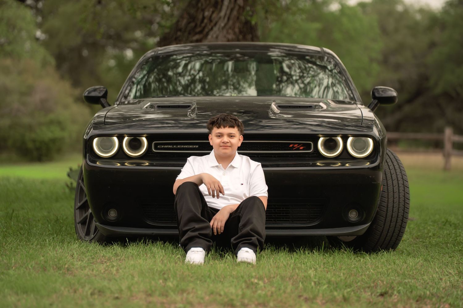 Young boy sits smiling in front of a black Dodge Challenger muscle car on green grass outdoors.
