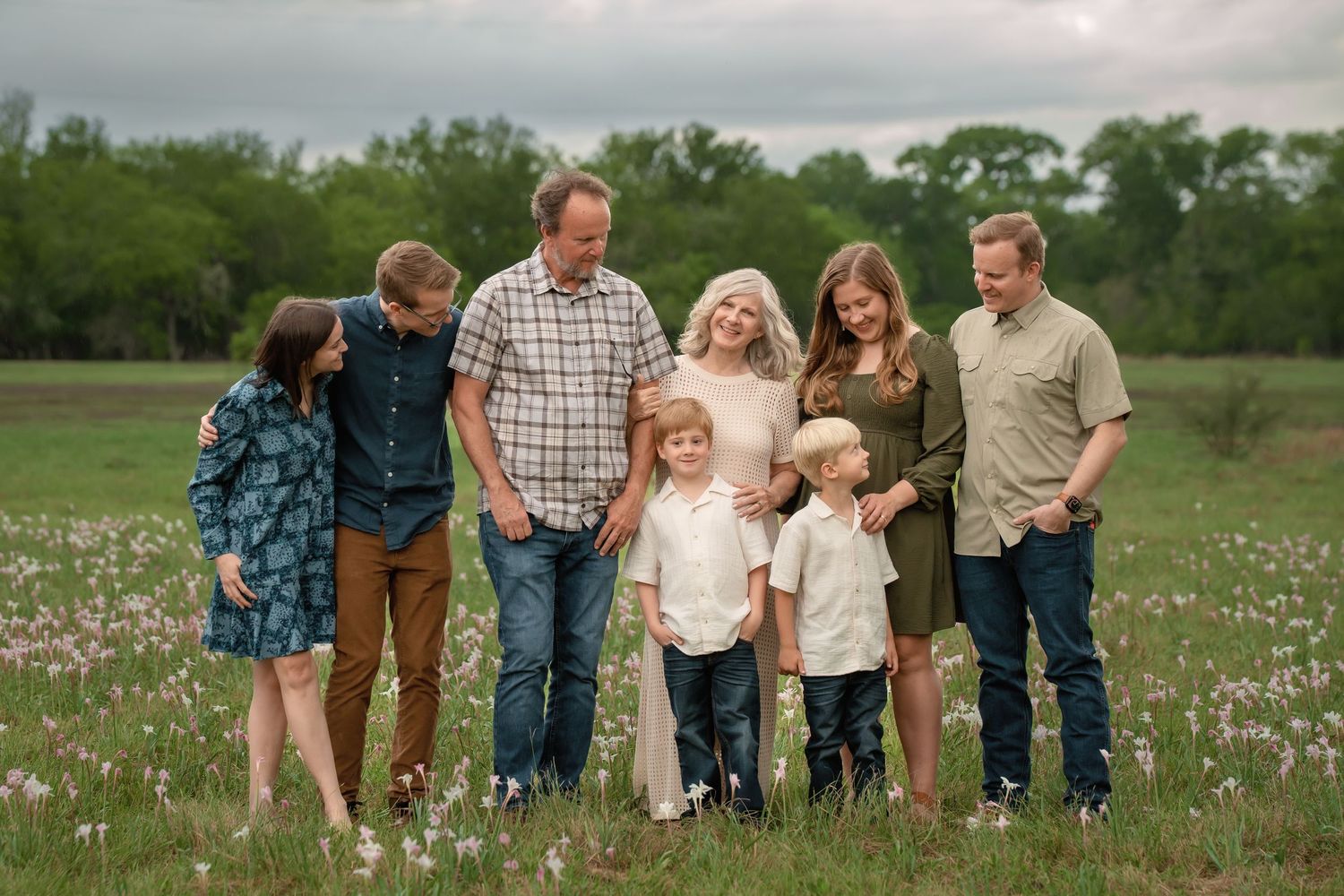 Family of seven posing together in a blooming wildflower meadow with lush green trees in the background.