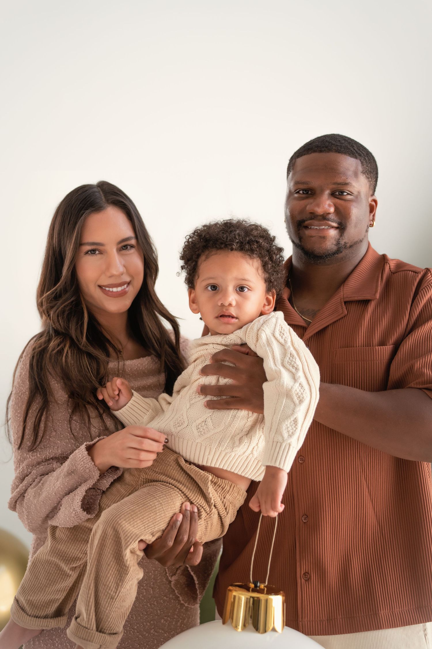A happy interracial family poses together, with a woman and man holding their young toddler in a warm, neutral-toned studio setting.