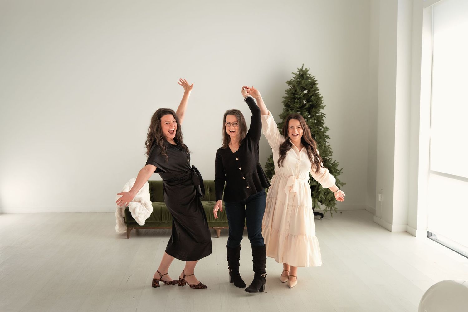 Three women dancing joyfully in a bright white room with a Christmas tree, wearing elegant black and white outfits.