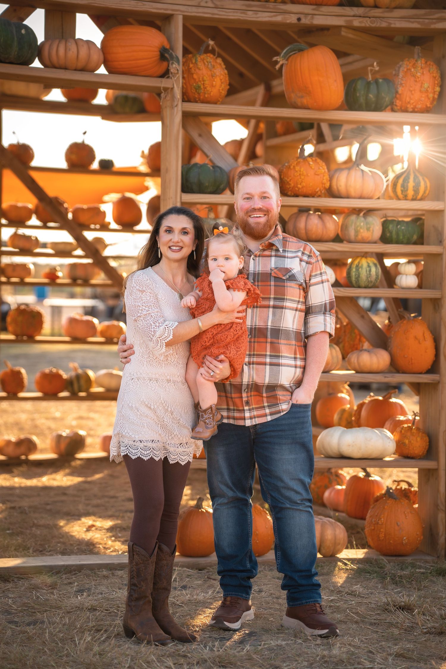 A family poses together at a rustic pumpkin patch display with wooden shelves holding autumn gourds and pumpkins.