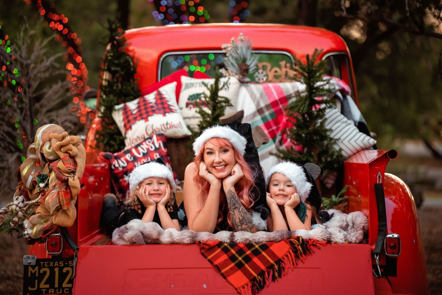 Three children in Santa hats pose joyfully in a decorated red vintage truck bed with Christmas trees and colorful lights.