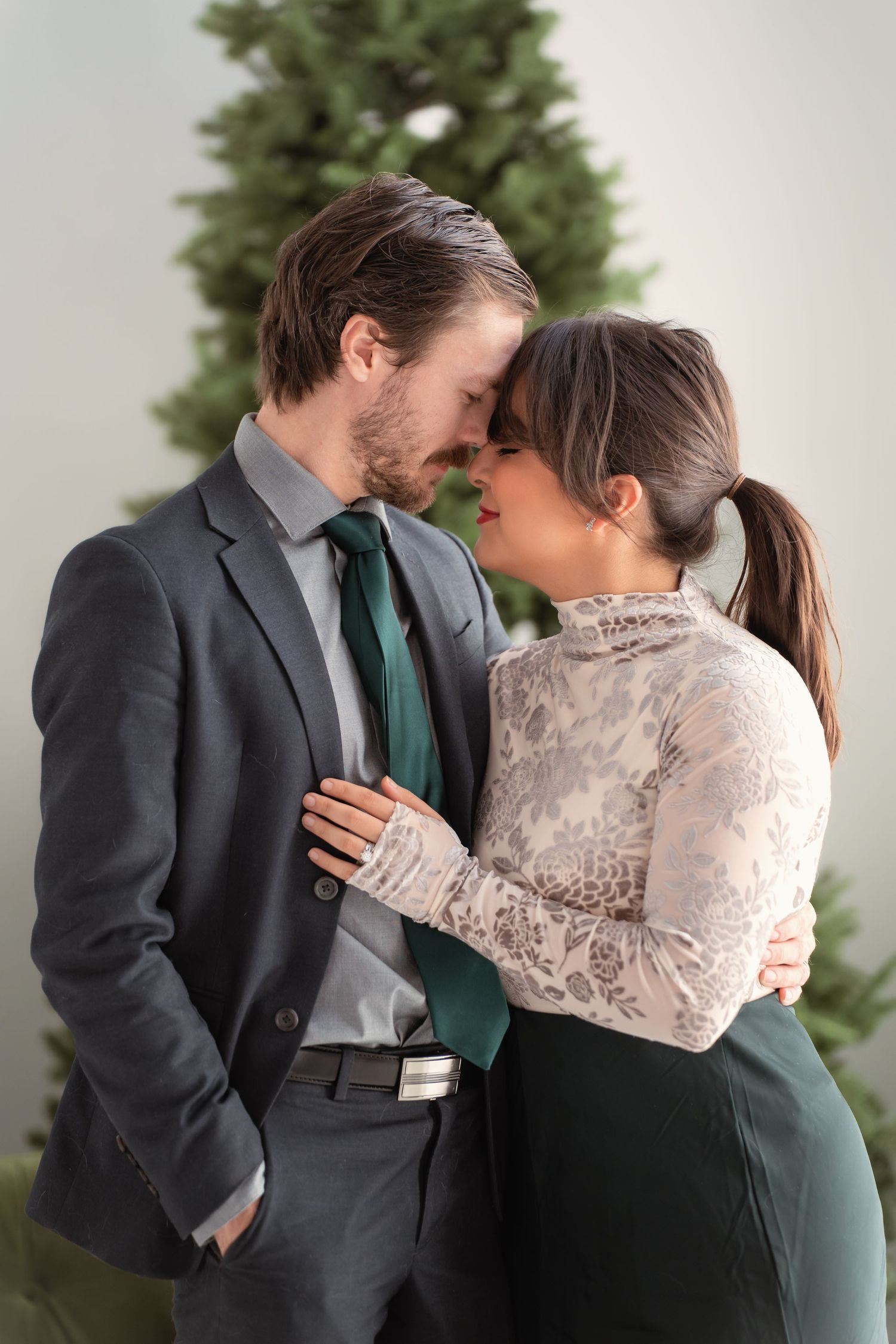 Romantic couple in formal attire sharing an intimate moment near a Christmas tree, woman in lace top embracing man in gray suit.