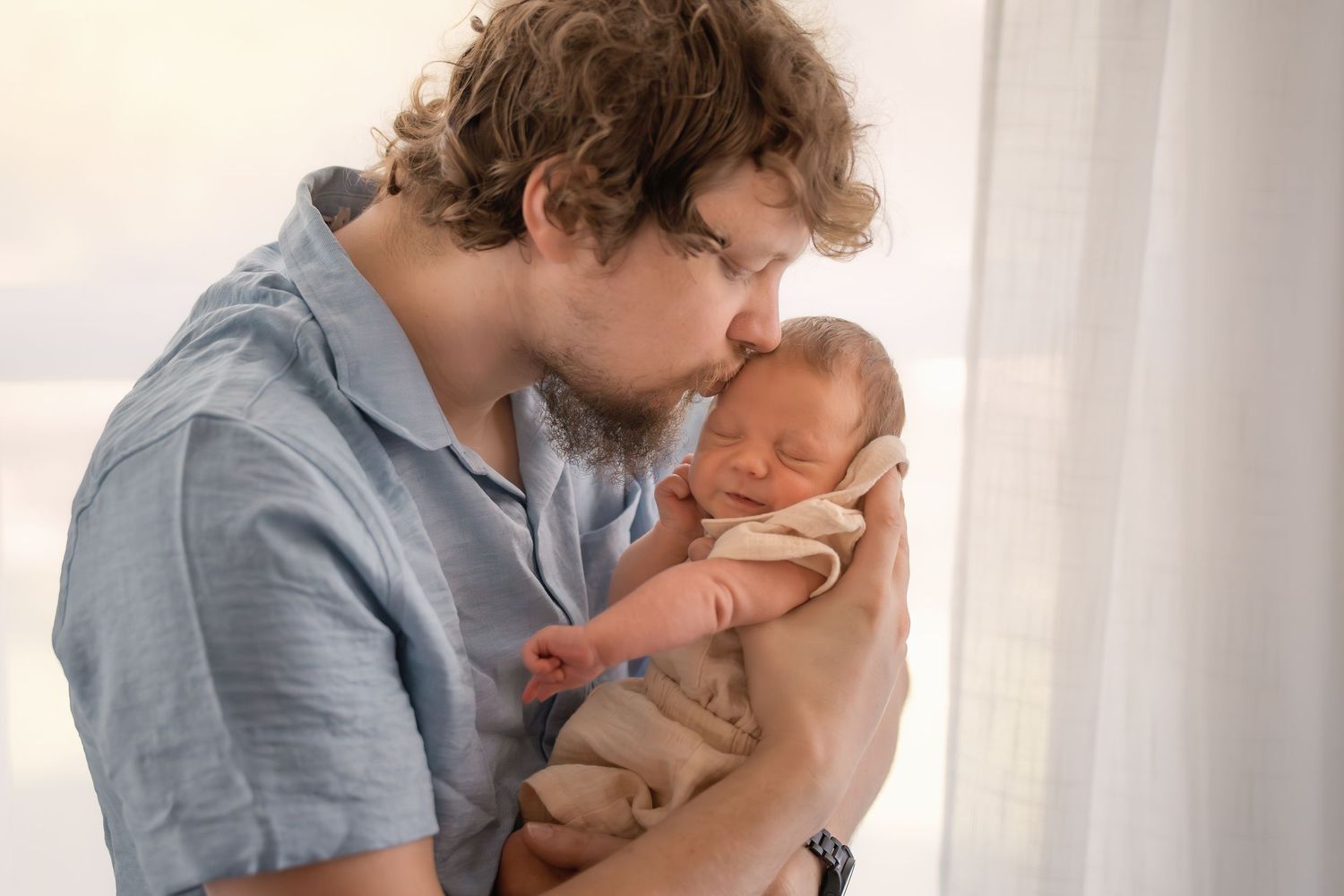 A father tenderly kisses his sleeping newborn baby wrapped in a tan blanket near a bright window.