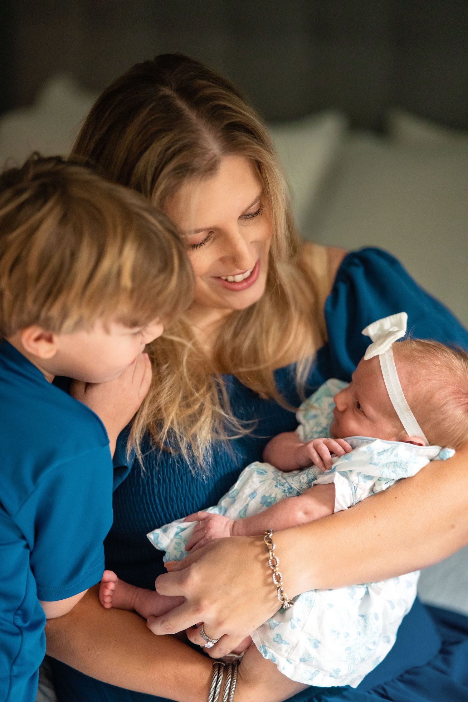 A smiling blonde mother in blue holds her newborn baby while an older child looks on tenderly.