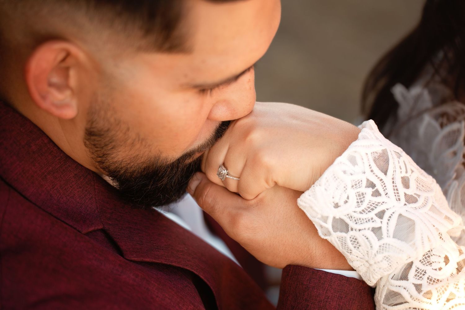 Groom in burgundy suit kissing bride's hand adorned with diamond engagement ring at wedding ceremony.