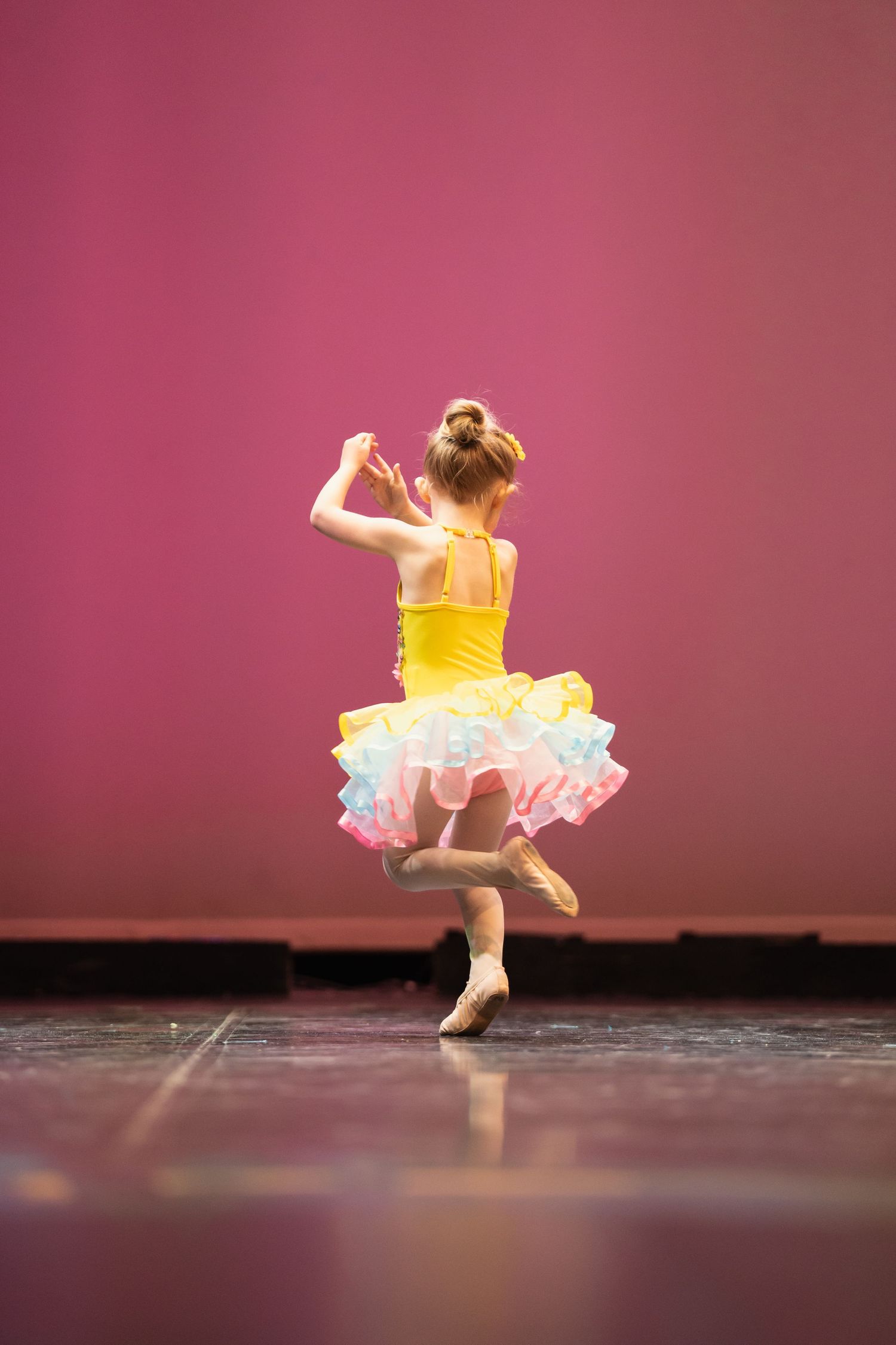 Young ballerina in yellow tutu performs on stage, dancing en pointe with arms raised against a pink backdrop.