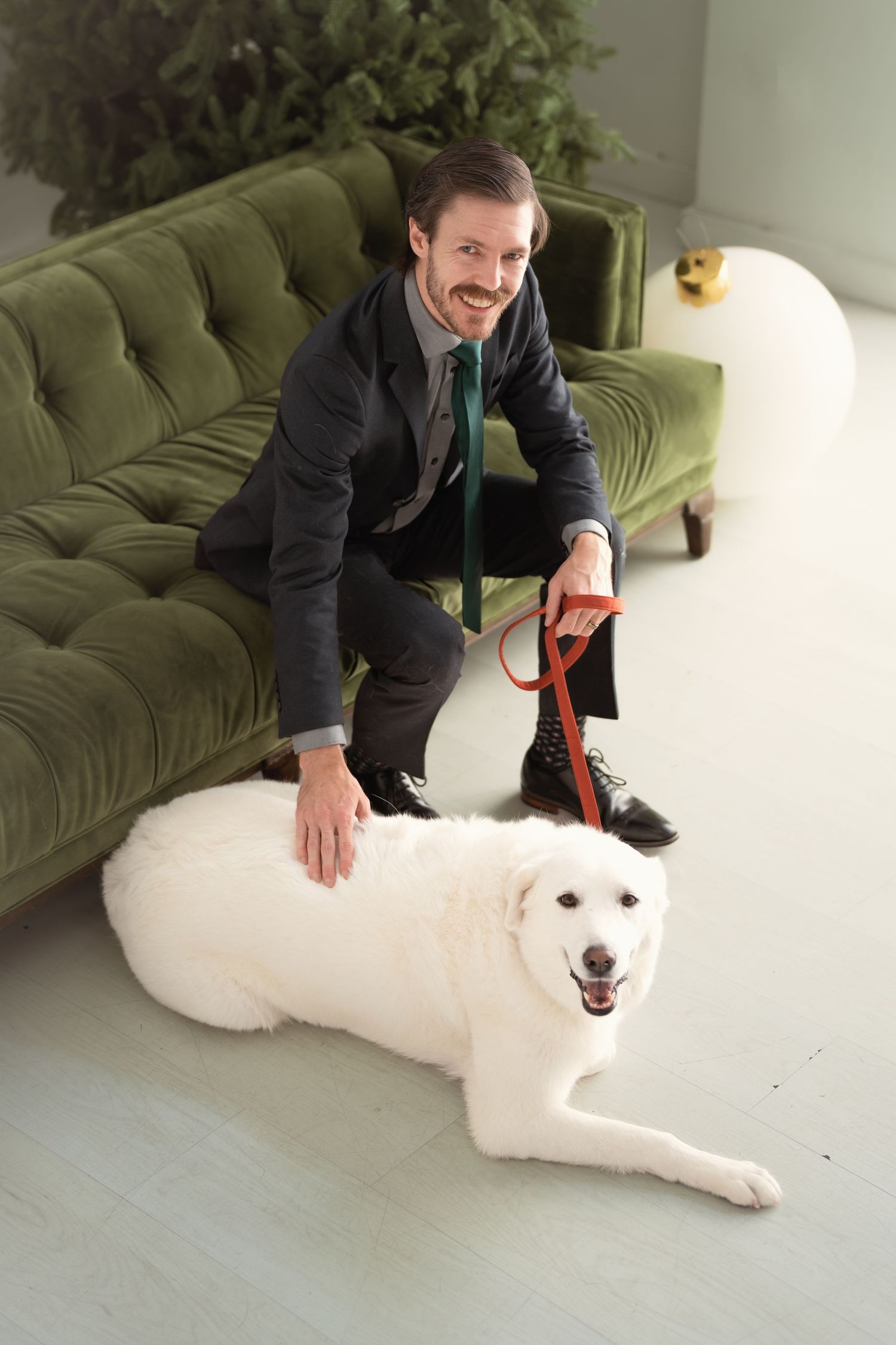A smiling man in a suit kneels beside a large white dog on a leash near a green velvet sofa indoors.