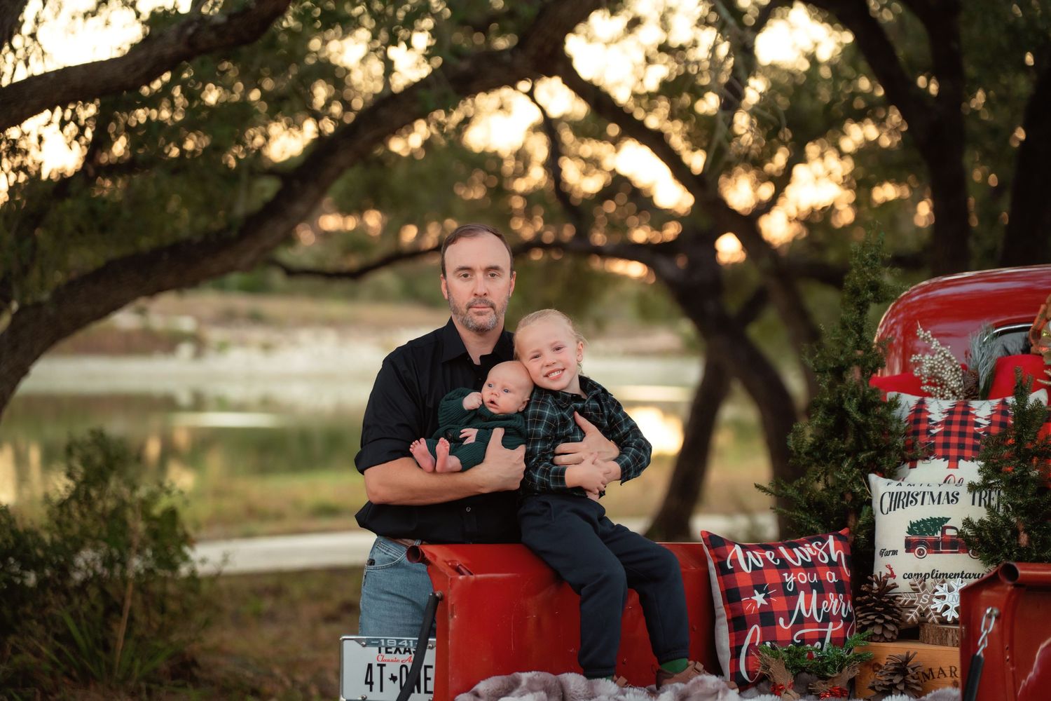 Man holding baby sits on red truck with Christmas decor, trees and golden sunset in background.